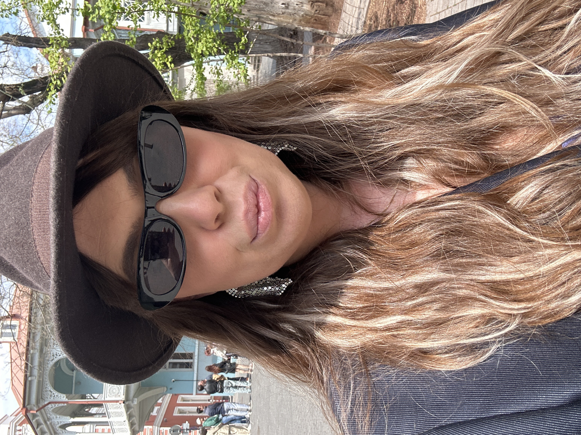 A close-up outdoor portrait captures a woman looking directly at the camera, her head slightly tilted. She has long, wavy hair with prominent blonde and light brown highlights, flowing over her shoulders. She is wearing large, black sunglasses that obscure her eyes, and a dark grey or brown fedora-style hat. Her lips are visible, appearing naturally pink with a subtle sheen, suggesting a faint smile. Large, shiny silver-toned earrings with a textured, chain-like design are visible on her right ear, peeking out from beneath her hair. Her attire includes a dark, possibly striped, top or jacket.
The background reveals an urban setting with architectural details. Parts of several buildings are visible, including one with ornate white decorative elements or balconies, and others with colorful facades (red and blue). Green foliage from trees with fresh leaves is seen in the upper background, suggesting spring or early summer. A bright sky indicates sunny weather and daytime. In the mid-ground, several people can be seen walking or standing, adding to the impression of a lively public space, consistent with a street scene in a city like Tbilisi, Georgia. There is no visible text in the image.