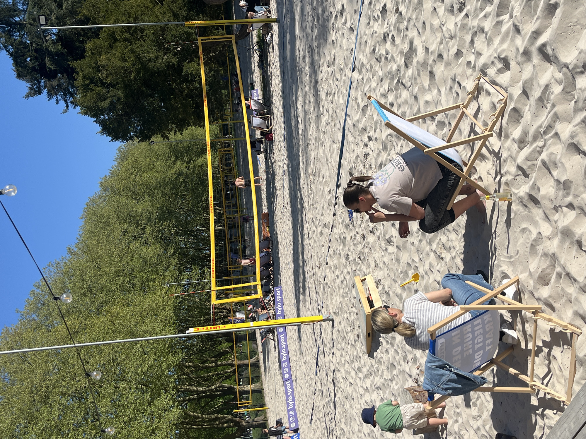 The image captures a sunny scene on a sandy beach volleyball court in Saarbrücken, Germany. Two women are sitting on wooden beach chairs on the right side of the frame, looking at something on the ground. One woman is wearing a gray t-shirt and black shorts, while the other is wearing a striped top and jeans. A small child in a green shirt and blue hat stands near the second woman's chair. In the background, a yellow volleyball net is visible, with trees lining the perimeter of the court. Other people can be seen in the background, some sitting in chairs, some playing volleyball. There are light bulbs strung above the court. The brand "Funtec" is displayed on the yellow volleyball net posts. The volleyball court also displays the brand "hylo.sport."