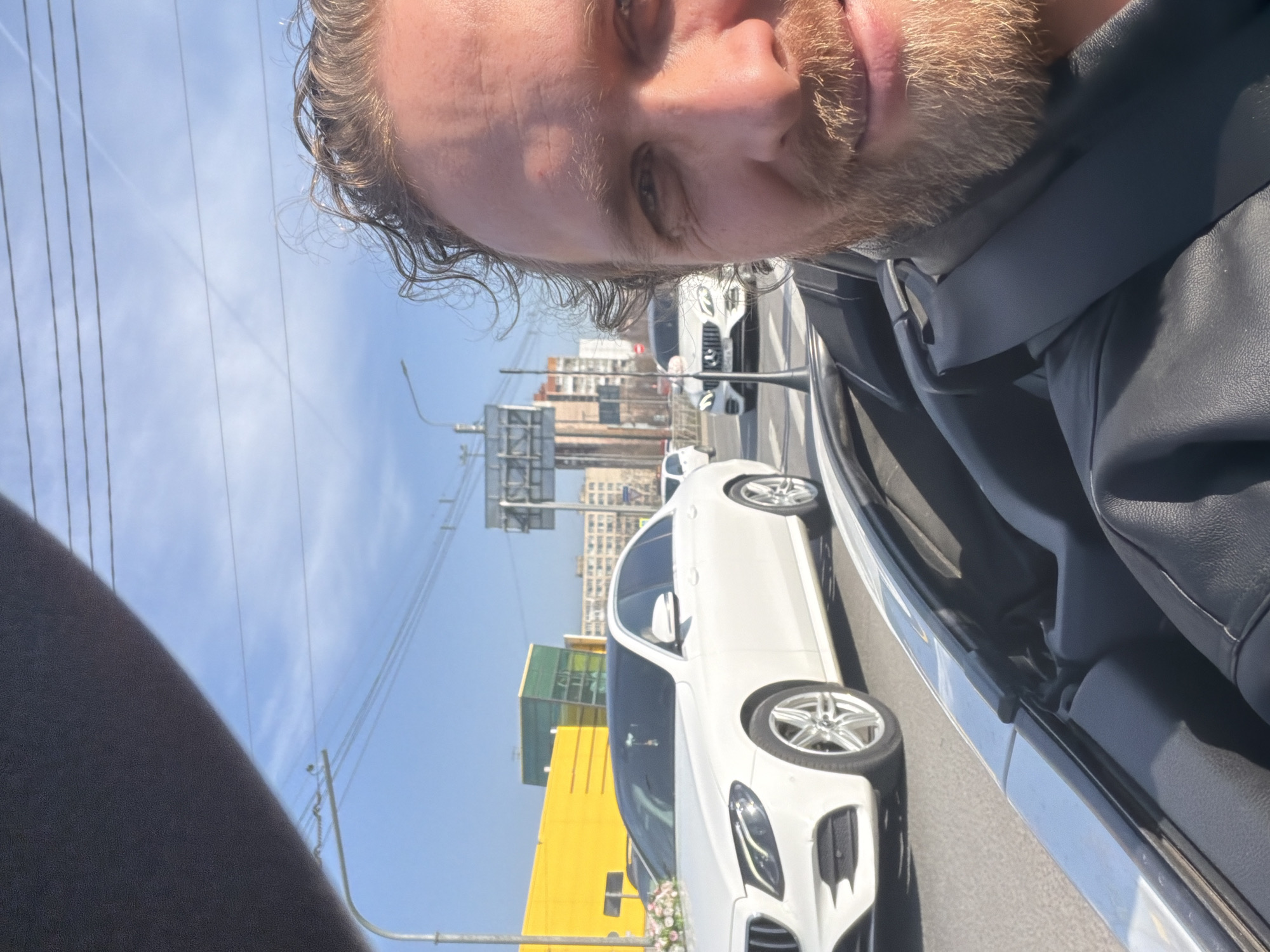 The image is a selfie of a light-skinned male with short, wavy blonde hair and a beard. He is wearing a dark-colored jacket. The picture is taken from inside a vehicle with the door open, revealing a white car parked on the street. Beyond the car, a street scene is visible with buildings, traffic lights, and overhead power lines. Based on the architecture and the signage visible, the location appears to be in an urban area. There is a yellow building visible across the road. There is a vehicle in front of the white vehicle. It is difficult to pinpoint the exact location in St. Petersburg without more context.