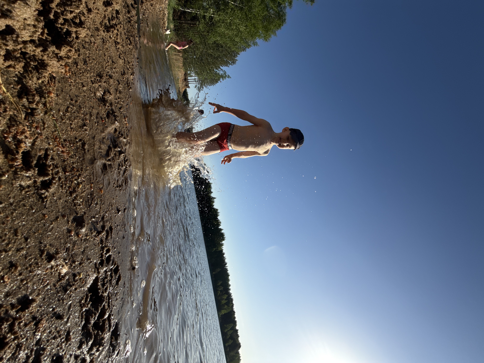 A young boy wearing red swim trunks is jumping into a body of water, creating a splash. The water appears to be shallow near the shoreline, which is sandy and textured. The location is Novyi, Russia. In the background, there are trees and another person wading in the water. The sky is blue and clear. The photograph has a low-angle perspective and is slightly rotated.