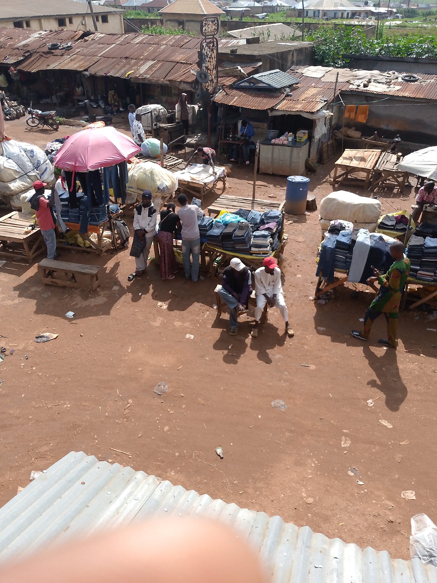 This aerial shot captures a bustling marketplace in Bukuru, Nigeria. The scene is dominated by stalls with corrugated iron roofs, some in disrepair, set against a backdrop of buildings and greenery. Pedestrians and vendors are engaged in commerce, with a variety of goods displayed, including clothing neatly folded on tables. A pink umbrella shades a collection of denim jeans. Some individuals are seated on benches, while others stand and interact with their surroundings, possibly examining merchandise or engaging in conversation. The ground is a dusty, reddish-brown earth, littered with scattered debris. A blue barrel and various bags and bundles add to the visual texture of the marketplace.