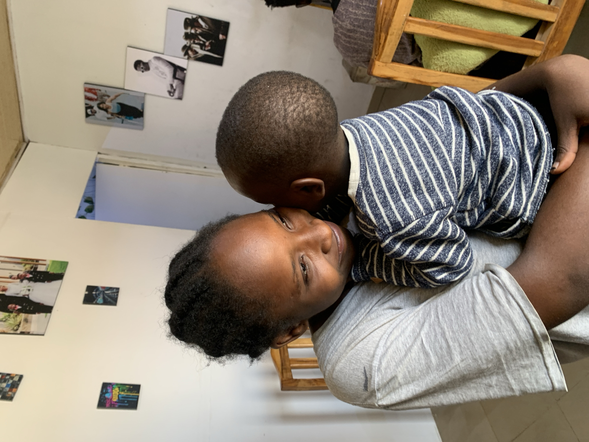 A woman is lying on her back, holding a young child against her chest. The child, who is wearing a blue and white striped shirt, is resting their head on the woman's face, near her ear. The woman is wearing a gray shirt. They appear to be indoors. On the wall behind them, there are several framed pictures. One picture shows a couple in a wedding attire, while others appear to be abstract images. A wooden chair is also visible in the background. It is not possible to confirm the location as Jos, Nigeria, from the visual information provided in the image.