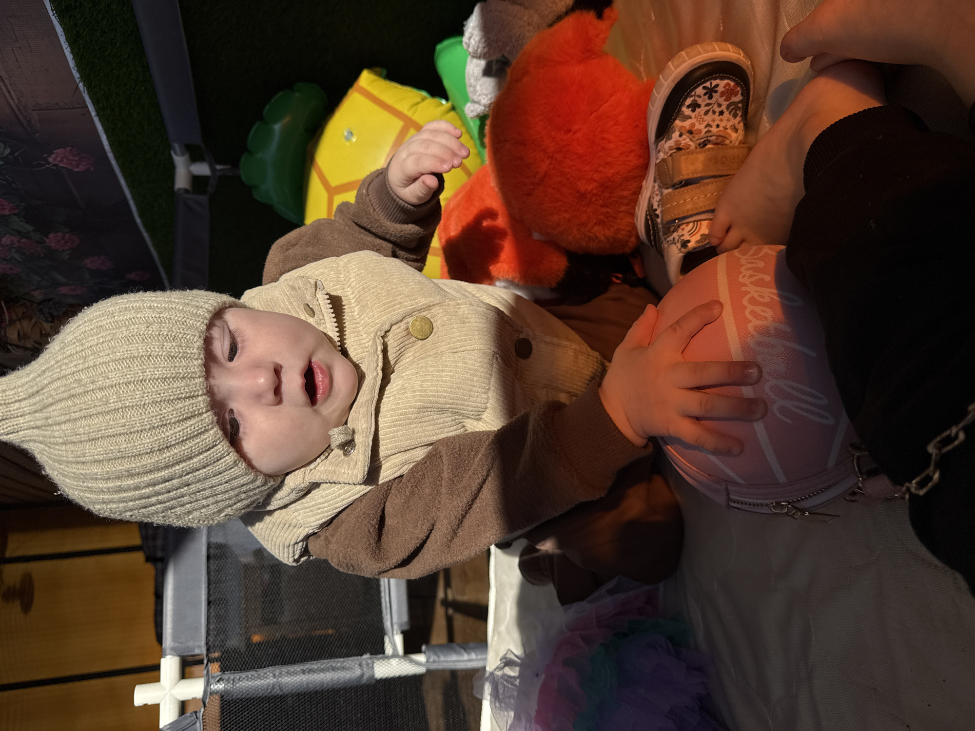 A young child wearing a beige knit hat and a brown and beige corduroy jacket is seated, facing the camera. The child is holding a pink basketball-shaped purse. Next to the child's foot, a small shoe with floral patterns is visible. Other items visible include a green and yellow inflatable toy, an orange stuffed animal, and a multi-colored tutu. The setting appears to be indoors, potentially in a play area, with a grey playpen structure visible in the background.
It is not possible to ascertain the location in the city of Кипчак, Russia from the provided image.