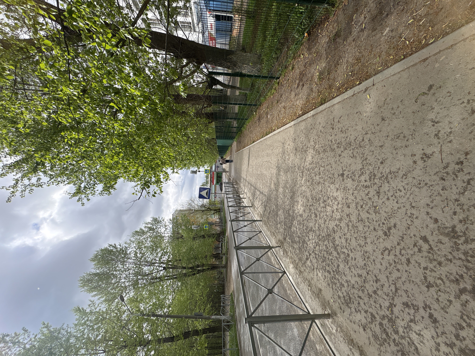 A sidewalk in Perm, Russia, is depicted with an oblique angle view. On the right side, a green wire fence separates the sidewalk from a strip of bare earth. On the left side, a metal fence runs alongside the sidewalk. Trees with lush green leaves overhang the sidewalk on both sides. In the distance, a pedestrian walks near a blue and white sign. Buildings are partially visible behind the trees and fences. The sky is overcast with grey clouds.