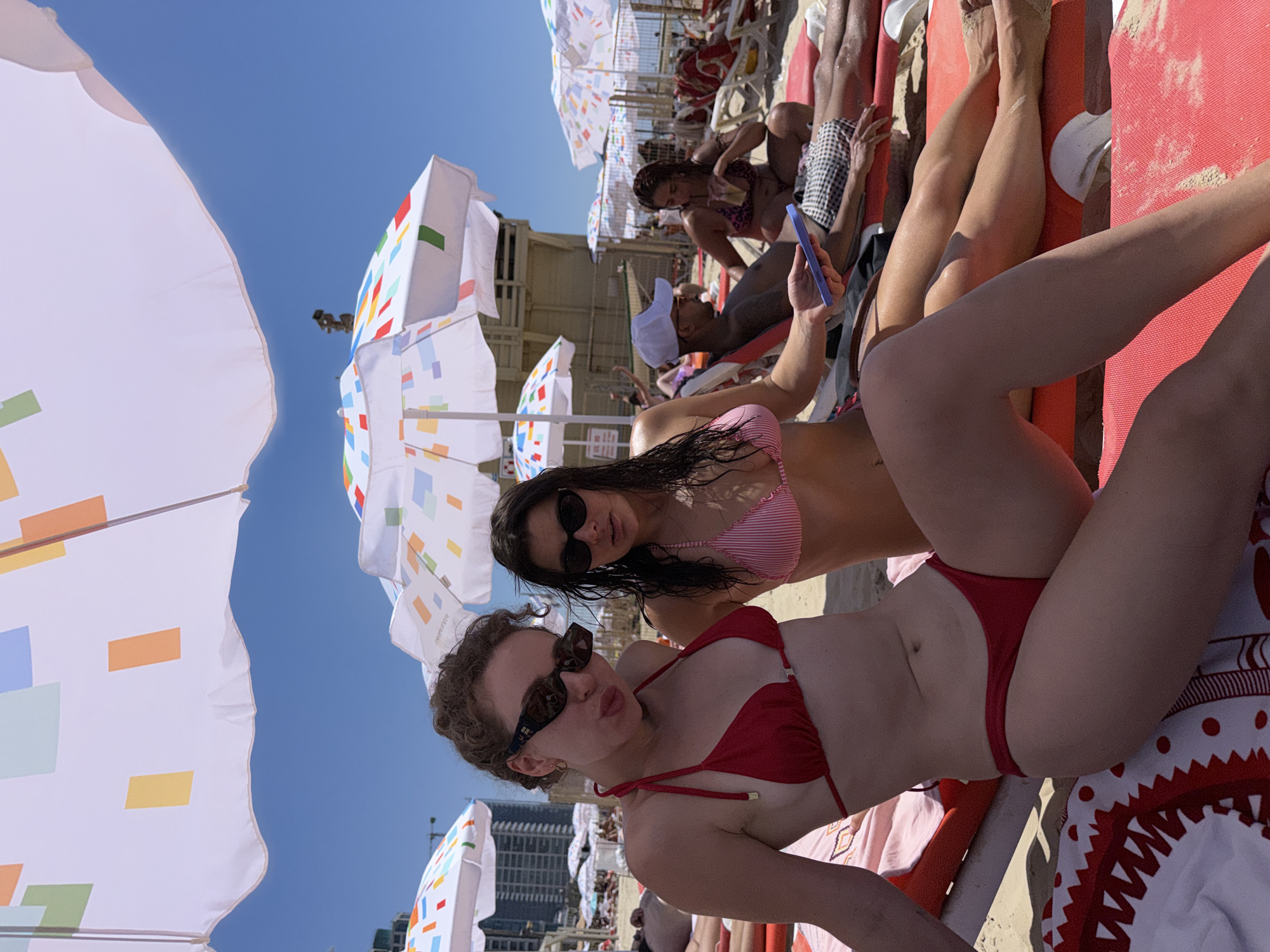 The image captures a sunny day at Gordon Beach in Tel Aviv, Israel. Two young women are sunbathing on orange beach chairs under a large umbrella with a colorful geometric pattern. The woman in the foreground wears a red bikini and sunglasses, with her curly hair framing her face. She is holding a blue smartphone. Beside her, a woman in a pink and white striped bikini also wears sunglasses. Behind them, other beachgoers are visible, along with beach umbrellas, chairs, and the sea in the background. A lifeguard station and high-rise buildings can be seen further down the beach. The overall atmosphere is relaxed and typical of a summer day at a popular urban beach.