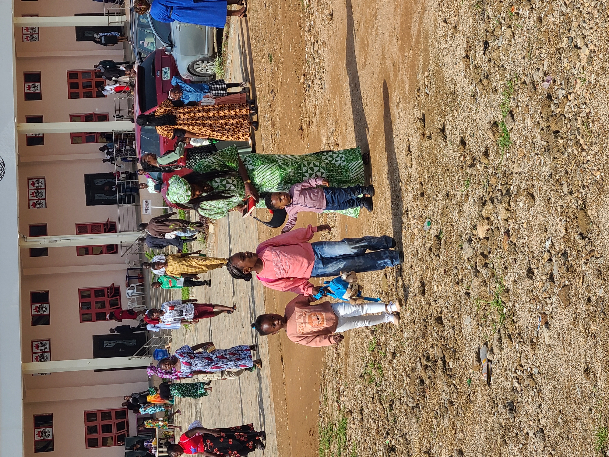 In Gura Top, Nigeria, a group of people are gathered outside a building with a light pink facade and red-trimmed windows. In the foreground, two young girls and a young boy are walking together on a dirt path. The older girl, on the right, is wearing a pink sweatshirt and blue jeans and is holding the hand of the younger girl, who is wearing a light brown sweatshirt and white pants. The boy, to the left of the girls, is wearing a light pink shirt and blue jeans. Behind them, a woman in a green and white patterned dress is holding a purse and what appears to be a red book. Several other individuals are visible in the background, some walking and some standing near cars parked on a paved area. The environment appears to be an outdoor setting with a mix of dirt and paved surfaces, with some sparse vegetation.