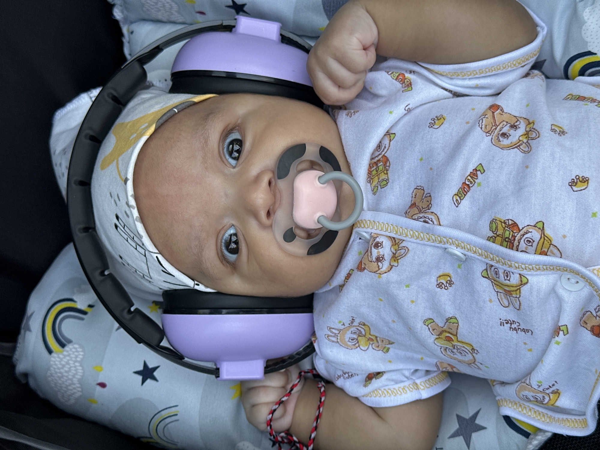 A close-up overhead shot shows a baby wearing lavender earmuffs and a pacifier, lying on its back. The baby is wearing a white onesie with a print of cartoon animals and the word "Labubu" repeated. The background consists of a soft, patterned fabric that appears to be part of a stroller or bassinet, featuring rainbows and stars. The lighting is natural, suggesting an outdoor or well-lit indoor environment. The baby's eyes are open and looking directly at the camera, and its expression is one of mild surprise or curiosity. A red, white, and black cord is visible wrapped around the baby's left wrist.