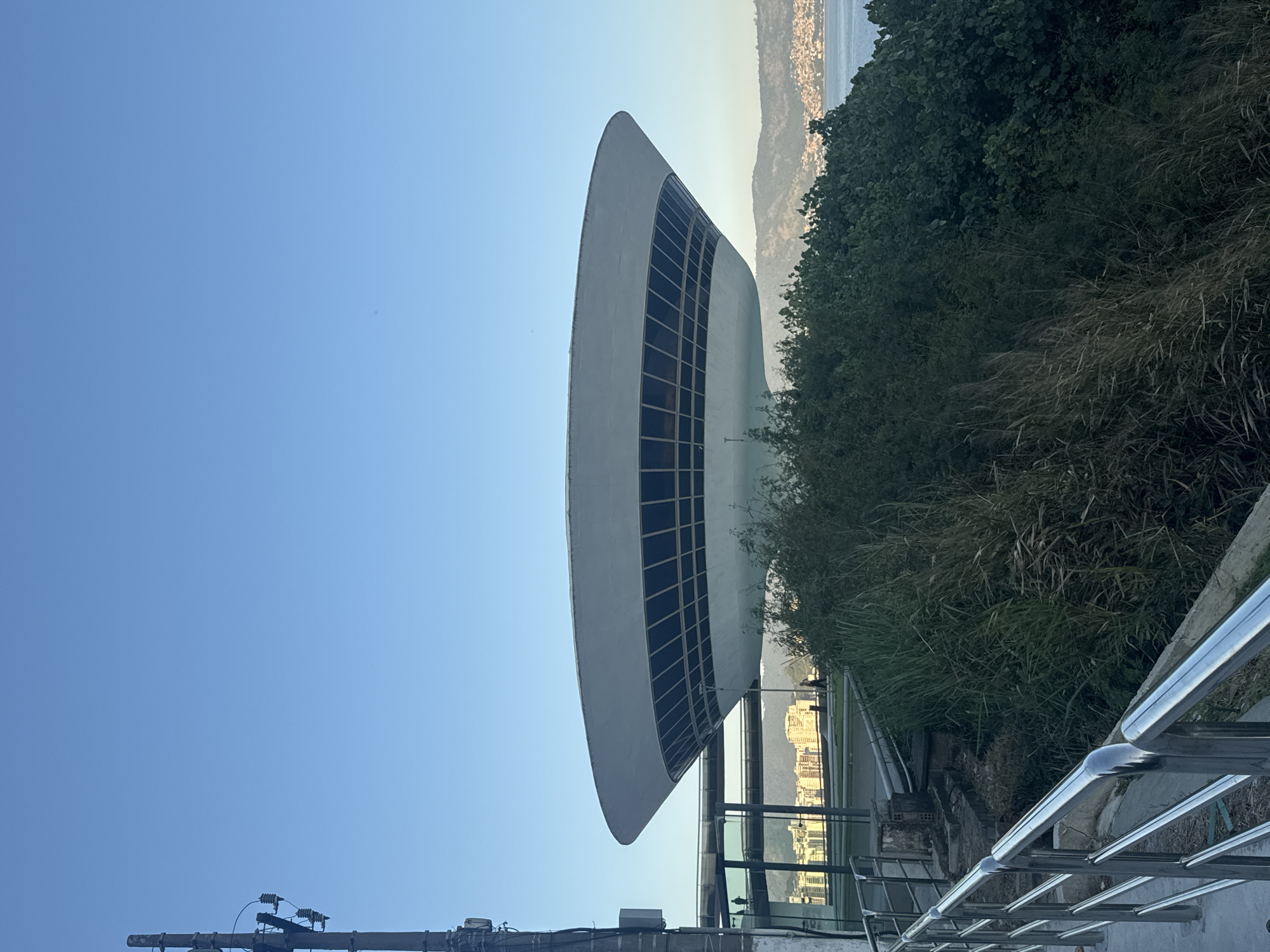 The image shows the Niterói Contemporary Art Museum (MAC) in Niterói, Brazil, with a clear blue sky above. The museum's distinctive saucer-like structure is prominently featured, with its circular form and band of windows. In the foreground, a metallic railing runs along a pathway, partially obscured by tall green grass and foliage. Beyond the museum, a body of water, likely Guanabara Bay, is visible, with distant hills and buildings forming the coastline of Rio de Janeiro on the other side. The scene appears to be captured during daylight hours, possibly late afternoon, given the soft lighting.