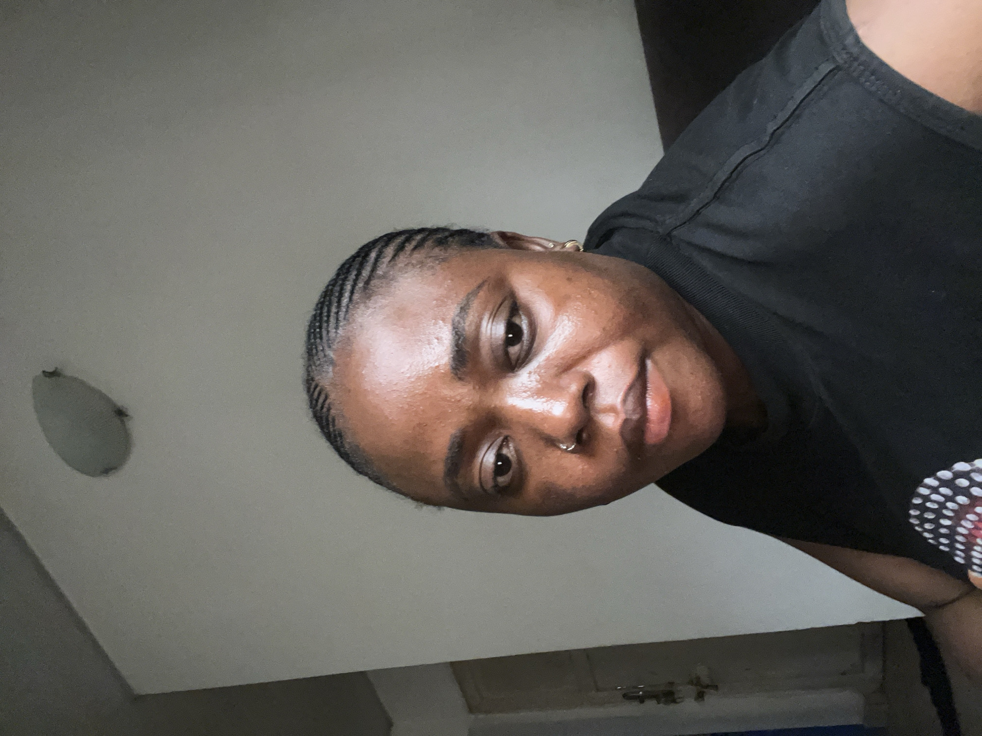 A medium shot taken from a low angle shows a Black woman with braided hair looking directly at the camera. She is wearing a black t-shirt and a gold hoop earring in her left ear. A small silver hoop is visible in her right nostril. The background consists of a white wall and a portion of a dark brown couch. A light fixture is visible on the ceiling. The image appears to have been taken indoors, possibly in a residential setting within the city of Abuja, Nigeria.