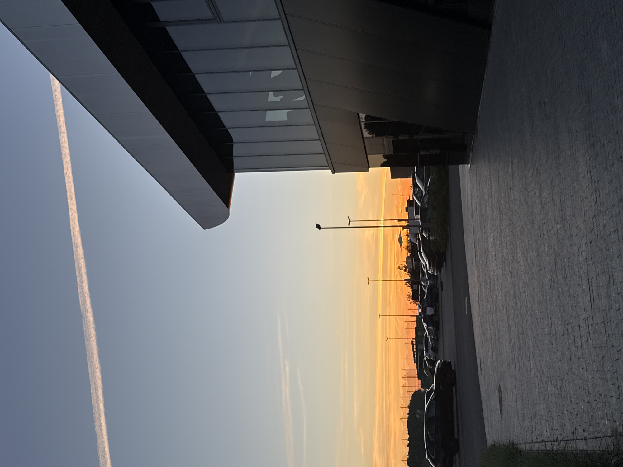 This image captures a sunset scene in Tallinn, Estonia, near the airport. In the foreground, a dark, modern building with large windows and a sleek, angled facade dominates the right side of the frame. The building's exterior is a mix of dark metallic panels and reflective glass, showing subtle reflections of the sky and interior lighting.
In front of the building, a paved parking lot is visible, with several cars parked in rows. The paving stones are dark and appear to be laid in a structured pattern. To the left of the parking lot, a grassy strip runs alongside the paved area, with some vegetation.
The background is characterized by a vibrant sunset sky. The horizon displays a spectrum of warm colors, ranging from deep orange to soft yellow, with streaks of pink and white from a distant contrail. Several tall light poles are scattered across the scene, and further in the distance, a sports field with netting and trees can be seen silhouetted against the colorful sky. The overall atmosphere is calm and serene, with the last rays of the sun casting long shadows.