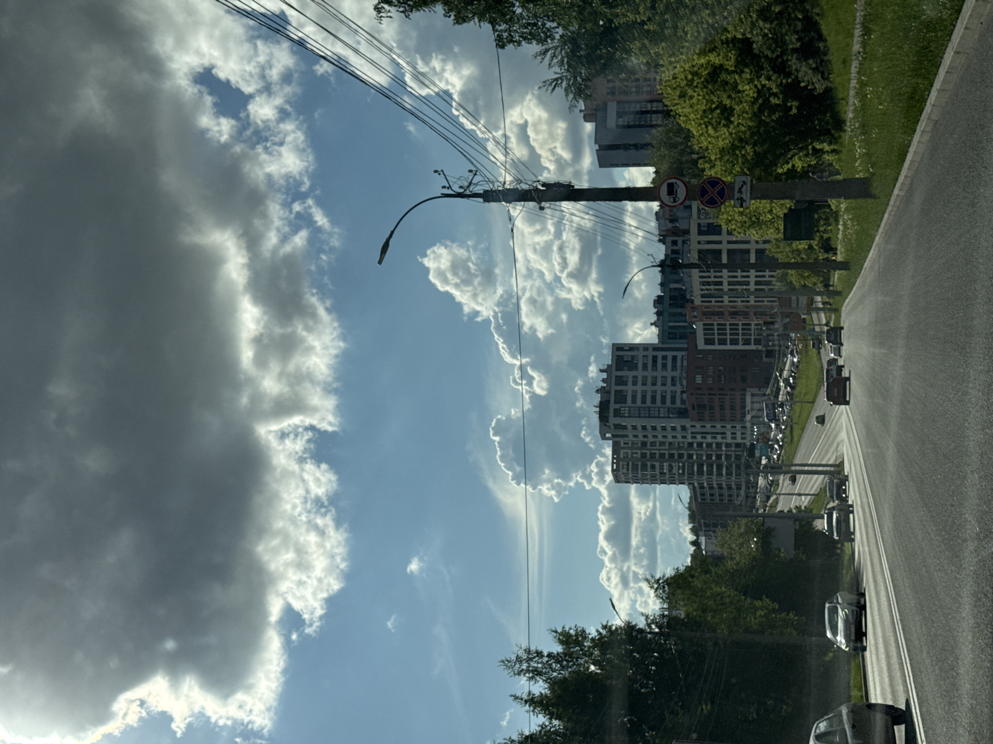 The image depicts a street scene in Izhevsk, Russia. The view is from inside a vehicle driving down the street. Tall, modern apartment buildings are visible on the left side of the road, interspersed with trees. There is a sidewalk with green vegetation alongside the buildings. The road appears to be asphalt, with white lane markings. Cars are present in the distance on the street. A utility pole with streetlights and multiple electrical wires runs along the left side of the street. The sky is predominantly blue, with a significant amount of puffy white and dark gray clouds. The light suggests a bright, sunny day. Road signs are visible on the right, near the apartment buildings.