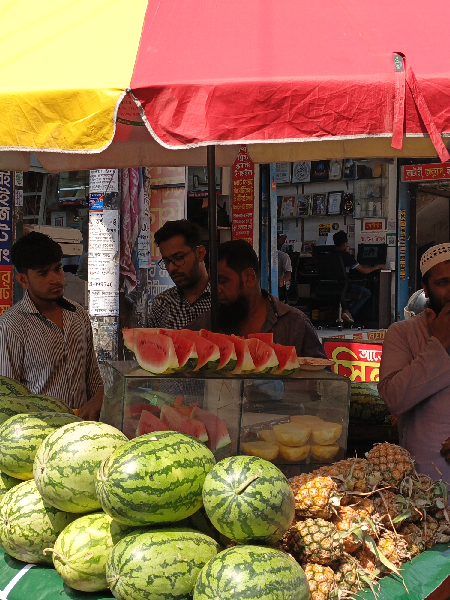 Here's a description of the image:
**Location:** This image appears to be taken at a bustling street-side fruit stall in Dhaka, Bangladesh, based on the signage in Bengali and the clothing of the people.
**People:**
* Four men are visible at the stall. One is wearing a striped shirt, another wears glasses and a checkered shirt, a third has a beard and wears a gray shirt, and a fourth man wearing a Muslim prayer cap.
**Subjects & Objects:**
* The fruit stall is filled with watermelons, sliced watermelon, pineapple, and small bowls of fruit.
* The stall is covered by a two-toned yellow and red umbrella.
* The background features posters and storefronts with signage in Bengali. A man is seated in the background behind a counter.
**Actions & Interactions:**
* It appears that the men are running the fruit stand, possibly selling fruit.
* One of the men wearing a prayer cap appears to be putting something in his mouth.
**Environment:**
* The scene is taken during the daytime under relatively bright sunlight, suggesting a busy and possibly crowded market street in Dhaka.