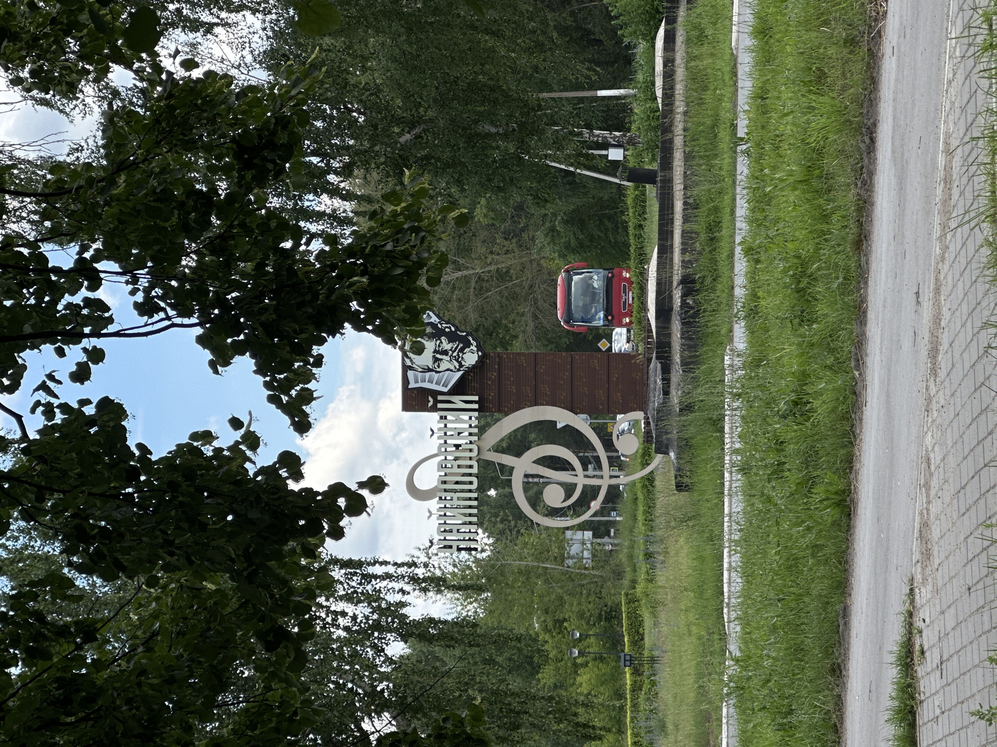 This is a landscape photograph taken outdoors on a sunny day. The scene features a roadside with a pathway and a grassy verge. In the background, there are trees and a red tractor. A prominent feature in the foreground is a large, decorative metal sculpture. The sculpture is a treble clef and appears to have text incorporated into its design, though it is difficult to read clearly. The text is white and vertically oriented, seemingly on a brown, musical-themed background. The overall impression is one of a public space, possibly a park or a community area, decorated with artistic elements related to music. The photograph is oriented vertically, with the sky and trees visible at the top and bottom, and the roadside and sculpture occupying the central and right portions of the frame. The weather appears to be clear and pleasant, judging by the bright light and blue sky with some clouds. The location is identified as Chaikovsky, Russia, which might suggest a connection to the composer Pyotr Ilyich Tchaikovsky.