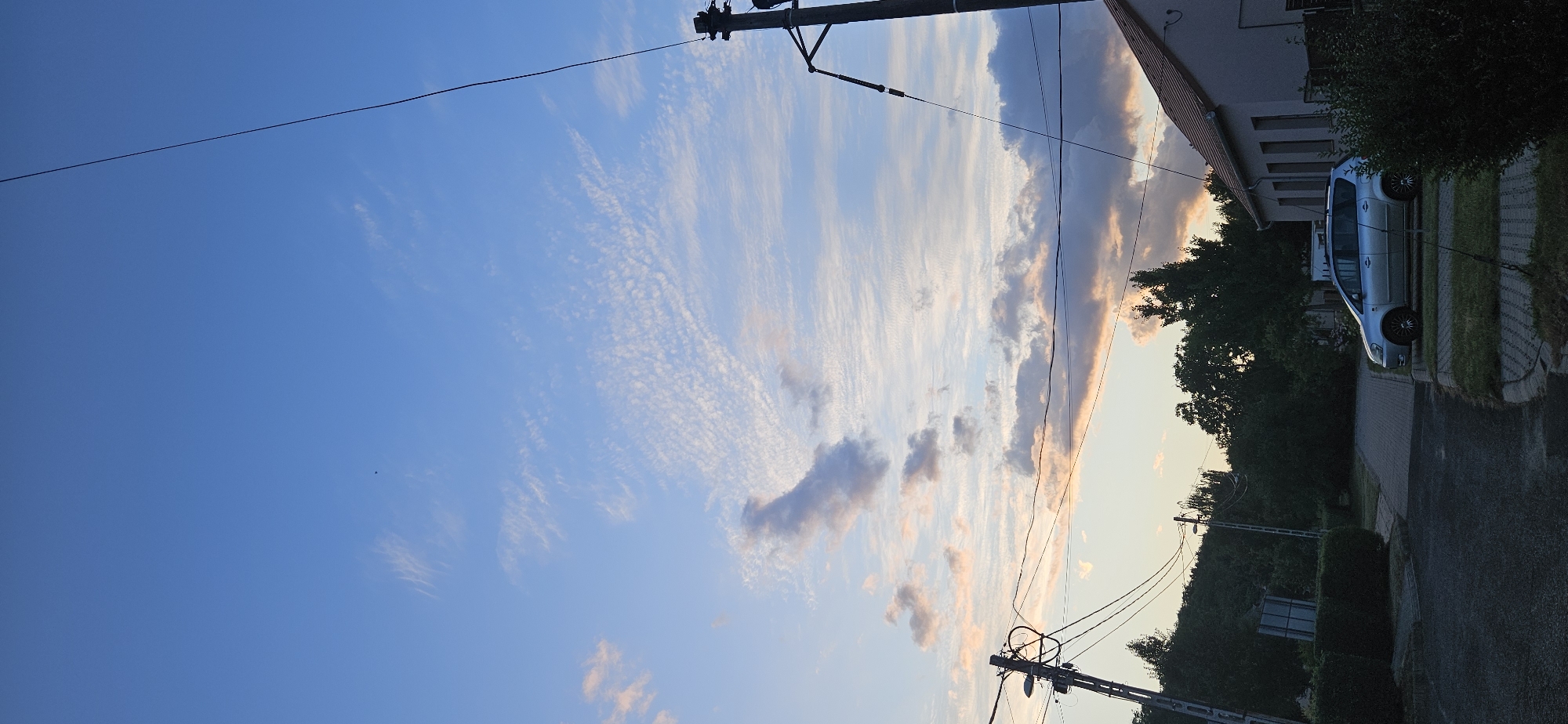 In the city of Hévíz, Hungary, a vertical, low-angle shot captures a serene evening sky transitioning from deep blue to lighter hues near the horizon, dotted with wispy, streaky clouds tinged with pink and orange. The sky is crisscrossed by utility wires. In the mid-ground, a line of dense green trees forms a natural backdrop. To the right, the upper portion of a light-colored building with a red-tiled roof is visible. In the foreground, a silver minivan is parked on a paved surface adjacent to a grassy area. Utility poles and more electrical wires are prominent throughout the scene, indicating a residential or semi-rural area.