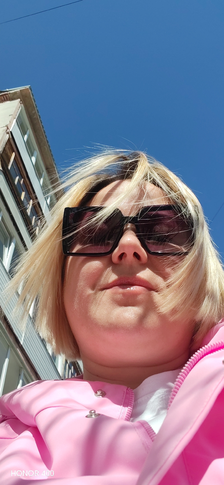 A low-angle, close-up photograph depicts a woman with fair, blonde hair and sunglasses wearing a pink jacket. The background includes a building with multiple windows on the left and a clear blue sky above. The bottom left corner of the image contains the text "HONOR 400". Based on the text, the photograph was likely taken with a mobile phone camera. The woman's hair is somewhat windswept.