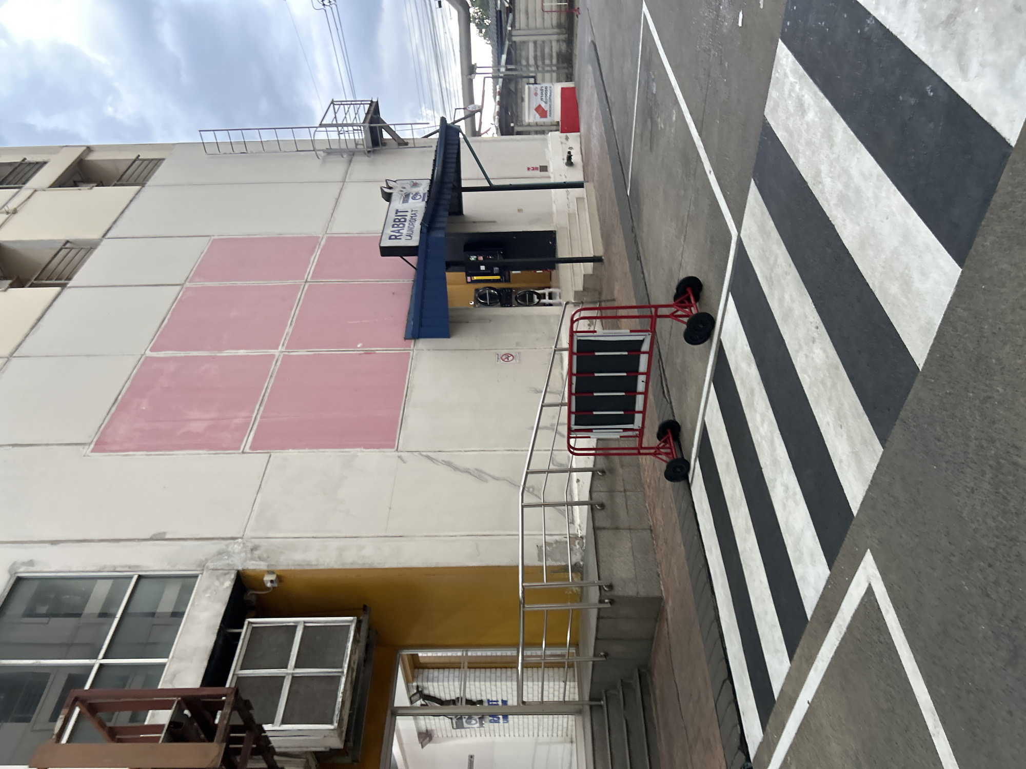 The image shows the exterior of a laundromat with a "RABBIT LAUNDROMAT" sign. The building is light-colored with large pink rectangular panels on its facade. A blue awning covers the entrance, where a set of stairs leads up to the door. Two washing machines are visible through a glass door to the left of the entrance. A metal fire escape ladder is attached to the side of the building. In front of the building, a black and white striped pedestrian crossing is painted on the asphalt. A red portable barrier with black panels is positioned on the pedestrian crossing. The scene appears to be in an urban or semi-urban environment, possibly a commercial area within Khlong Luang Town Municipality, Thailand, given the visual cues and common urban development in such areas. The sky is cloudy.