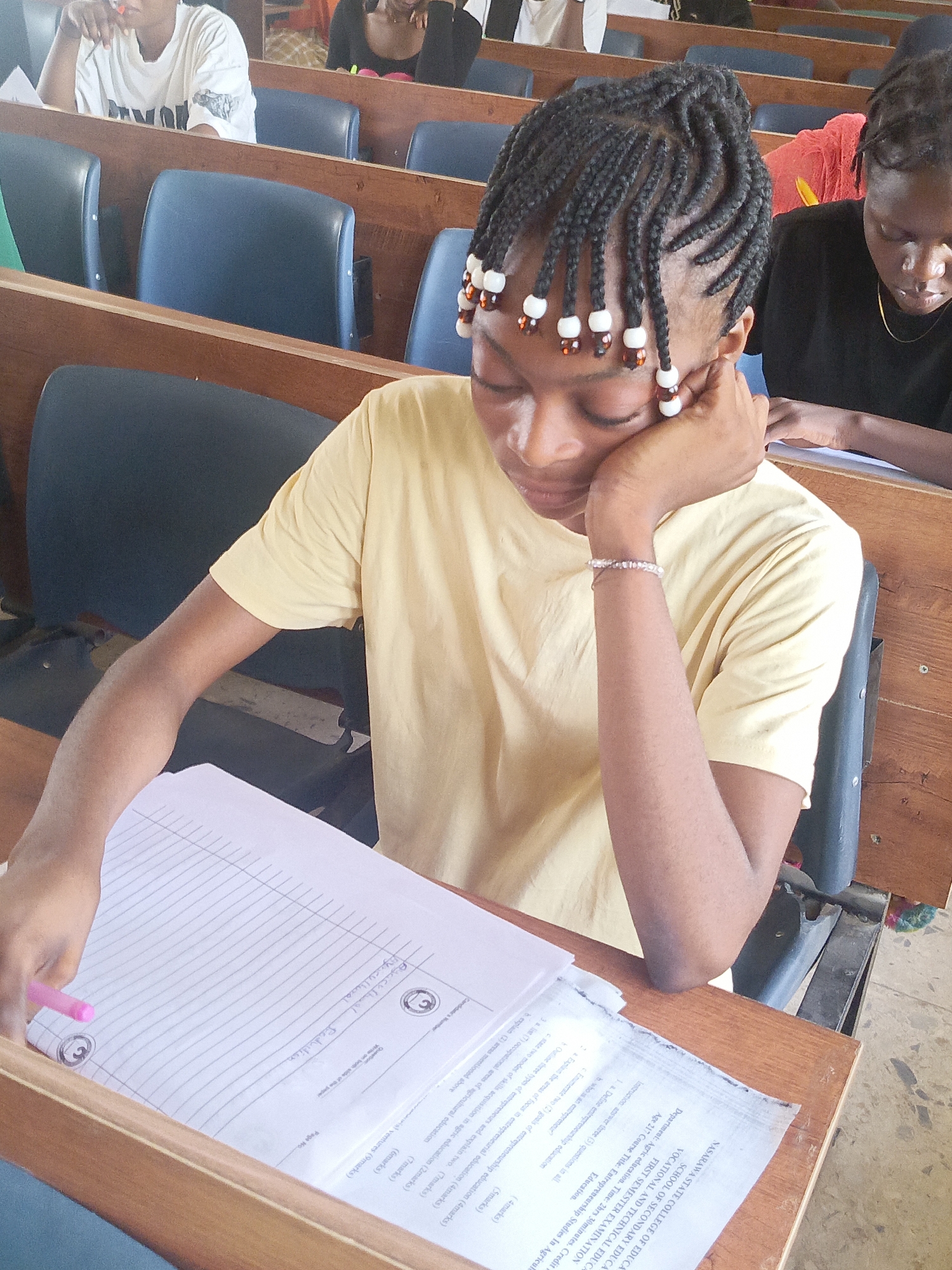 In Akwanga, Nigeria, an image captures a student in a classroom setting. The young woman, adorned with braided hair featuring decorative beads, is seated at a wooden desk. She is wearing a yellow t-shirt and a bracelet. Her left hand rests on her cheek as she appears to be engaged in or contemplating the exam paper in front of her, which is titled "Entrepreneurship Studies in Agricultural Education". On the desk are two papers, an exam question paper, and an answer booklet where the student has written "Viyicultural" and "Asciculhwal Production". The student is holding a pink pen in her right hand. Other students can be seen seated in rows of blue chairs with wooden desks, suggesting an examination or lecture in progress. The classroom lighting appears bright.