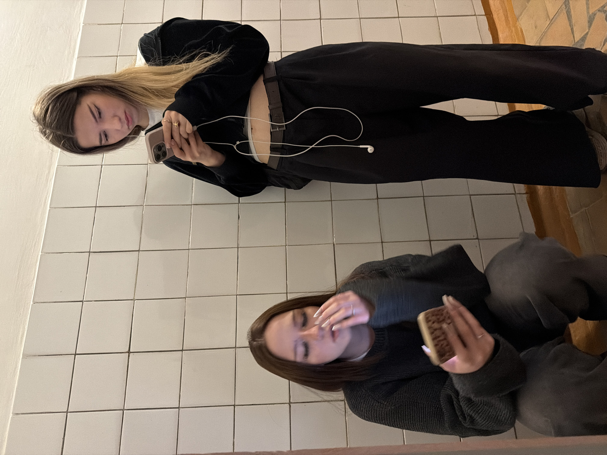 The rotated image displays two young women indoors, likely in Noviy, Russia, against a background of white square tiles.
In the foreground (upper section as rotated), a woman with long, light brown hair with blonde highlights is seen looking down at a light pink/beige smartphone held in her hands. White wired earbuds are connected to the phone and rest on her abdomen. She wears a dark long-sleeved top, black trousers, and a dark belt over a light-colored undershirt, which is visible above her midriff. Rings are visible on her right hand. Her expression is focused or pensive.
Further back (lower section as rotated), another woman with long, straight dark brown hair is also looking down, holding a smartphone with a textured, possibly brown and cream, case. She wears a dark grey or black knitted sweater and dark trousers. A ring is visible on her left hand, and she has long, light-colored fingernails. Her gaze is directed at her phone.
The setting appears to be an interior space with white tiled walls or floor. Dark grout lines segment the tiles. To the far right, a strip of warmer-toned material, possibly wood or different tiling, borders the white tiles. The lighting is uniform, suggesting an indoor environment. Both individuals are absorbed in their smartphones, creating a sense of individual focus within a shared space. No visible text is discernible.