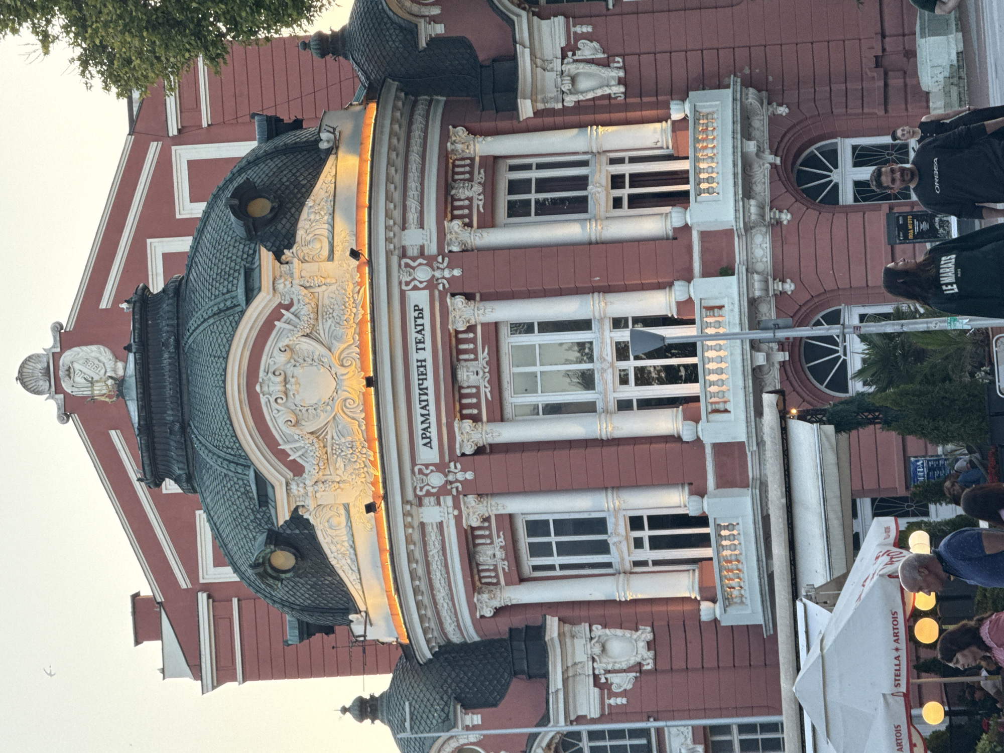 A low-angle, close-up shot captures the ornate facade of a large, salmon-pink building, likely a theatre, in an urban setting. The building features a dark green, domed roof and intricate white detailing, including columns, balconies, and decorative sculptures. The name of the building, "ДРАМАТИЧЕН ТЕАТЪР" (Dramatic Theatre), is visible in white lettering on the facade. In the foreground, patrons are seen dining or relaxing outdoors under white umbrellas advertising "STELLA + ARTOIS." A traffic light pole stands to the left of the frame. The sky is a soft, hazy yellow, suggesting either dawn or dusk. The overall atmosphere is one of architectural grandeur and relaxed outdoor leisure.