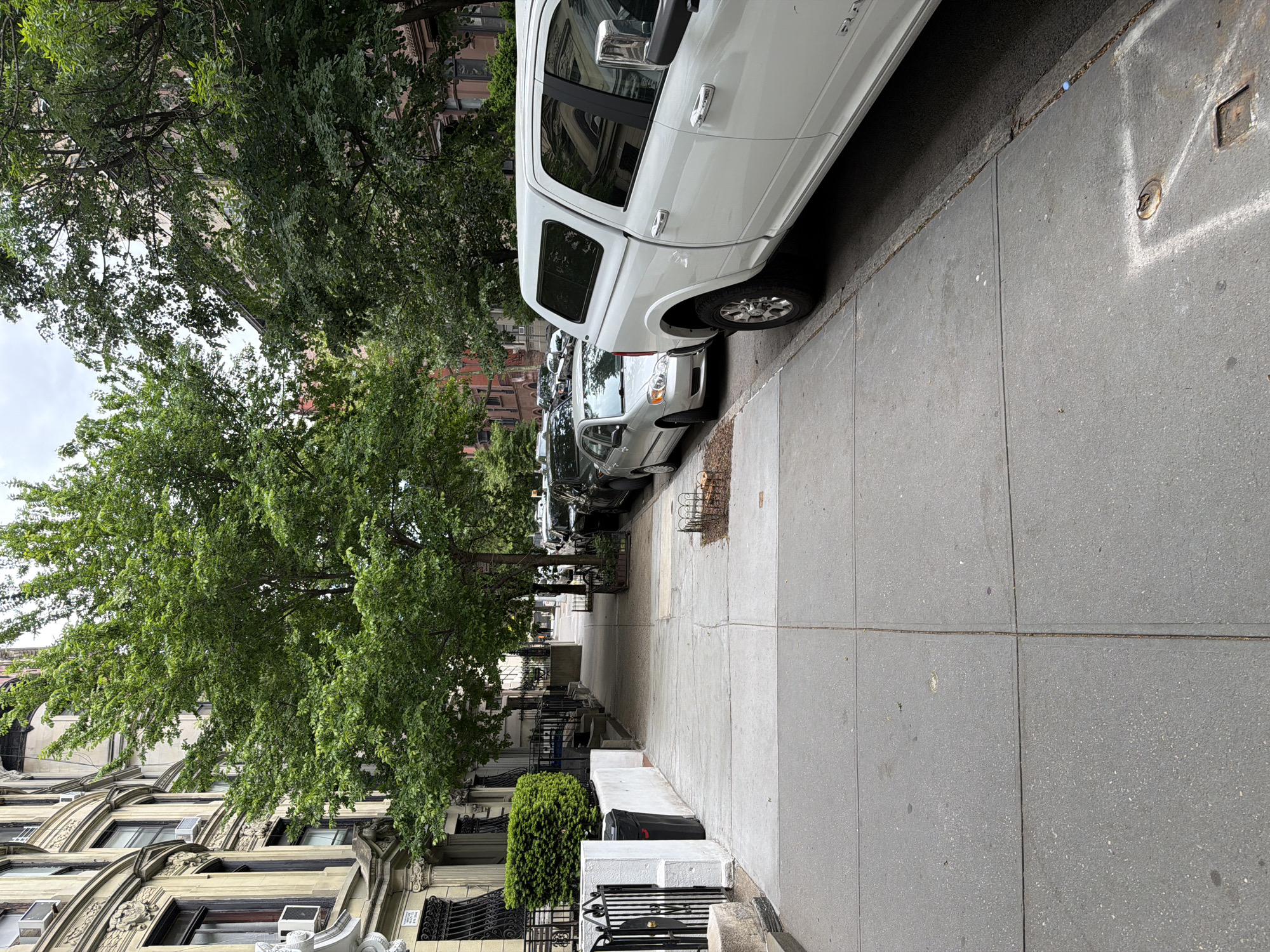 The image depicts a street scene in a residential area of New York City. The focus is on a tree-lined sidewalk with parked cars along the curb. Row houses with ornate architectural details are visible on the left side of the street. A large white truck and other vehicles are parked along the right side of the street. The sidewalk is paved with grey concrete. A small green bush and wrought iron fencing are present in front of one of the houses. The sky is overcast. The scene is still and there are no visible people.