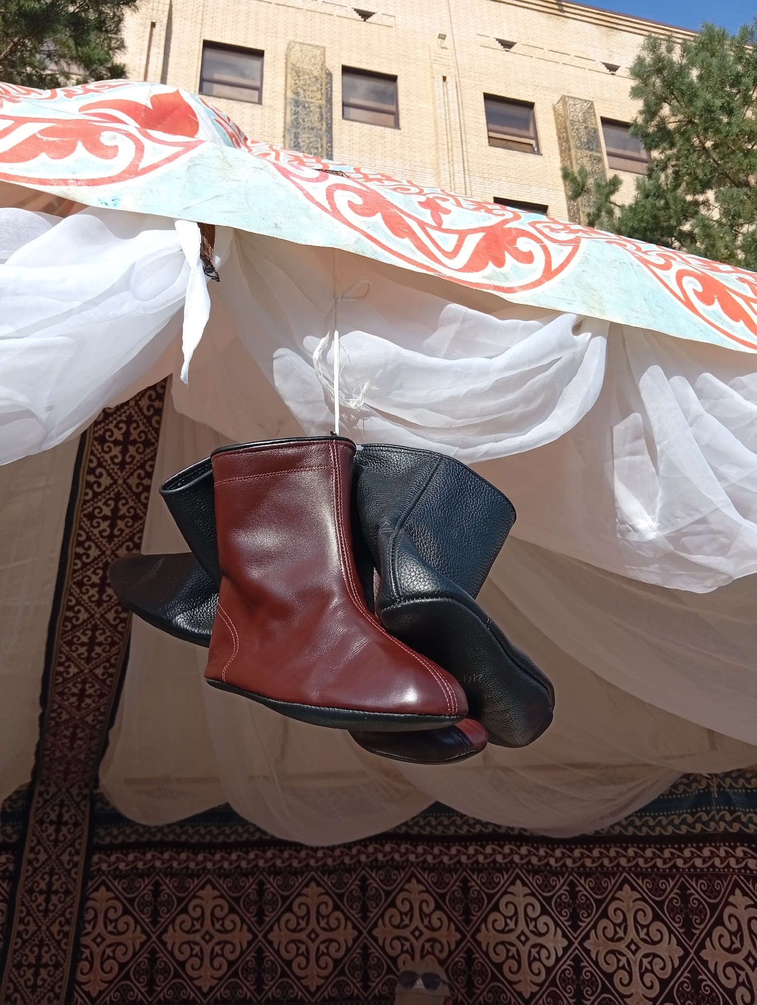 The image shows a close-up of two pairs of boots hanging from a string. One pair is a dark, rich brown color, and the other is black. Both pairs appear to be made of leather and are of a low-cut boot style. They are suspended in front of a white, sheer fabric, and behind that, a patterned tapestry or rug with intricate designs in brown and beige is visible. The background consists of a light-colored brick building with multiple windows, some of which have dark frames. Green foliage from trees is also present, and the sky is a clear blue. The overall scene suggests an outdoor market or display, possibly a cultural event, given the traditional-looking tapestry. This scene is located in the city of Kostanay, Kazakhstan.
