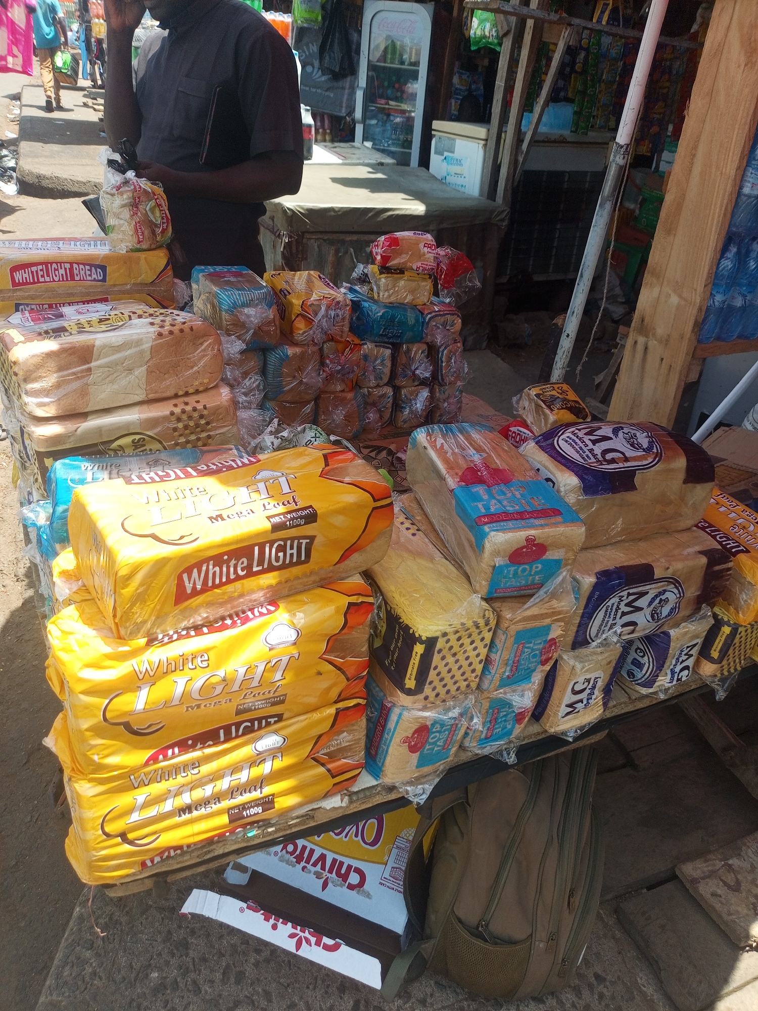 This image captures a vibrant outdoor market scene, likely in Nigeria, characterized by an abundance of bread for sale during bright daylight hours. The central subject is a market stall piled high with numerous loaves of packaged bread. A man, dressed in a dark short-sleeved shirt, is partially visible behind the stall, appearing to attend to the goods or possibly engaging in a phone call. In the background, other individuals are seen walking, and makeshift market structures and shelves are discernible, containing various other products like bottled drinks and coolers.
The bread on display features several brands and packaging styles. Most prominent are the "White LIGHT" brand loaves, easily identifiable by their bright yellow packaging, frequently labeled "Mega Loaf NET WEIGHT: 1100g." Other brands include "TOP TASTE MODERN BAKERY" with blue and white packaging, and "MG Modern Bakery" loaves distinguished by purple and white wrapping, some marked "Bromate Free." Many loaves are individually wrapped in clear plastic. Underneath the display table, a beige backpack sits on the ground next to a cardboard box.
Visible text on the bread packaging includes "WHITELIGHT BREAD," "White LIGHT Mega Loaf NET WEIGHT: 1100g," "TOP TASTE MODERN BAKERY," "MODERN BAKE," "MG Modern Bakery," and "Bromate Free." A partially visible box underneath the stall shows text like "Chivito" and "HANDLE WITH CARE." A "Coca-Cola" cooler is visible in the background. The scene suggests a busy local commercial area focused on daily provisions.