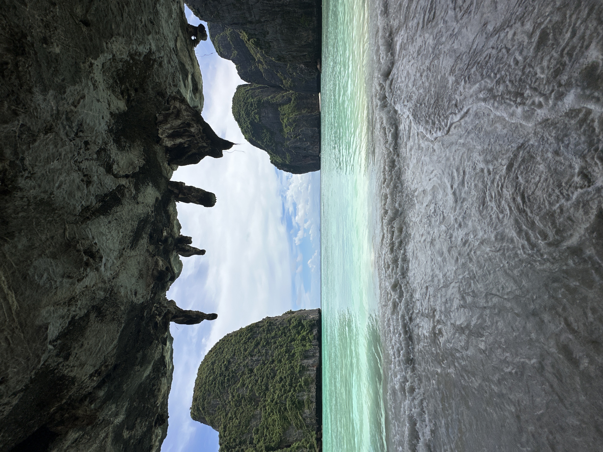 This is a medium-wide shot of a beach scene viewed from inside a cave opening. The cave ceiling, composed of rough, dark rock with some lighter, textured patches, hangs low and obstructs the top portion of the frame. Several stalactites, or rock formations resembling icicles, hang from the cave's underside.
Beyond the cave opening, a bright, aquamarine ocean stretches out. Gentle waves with white foam are breaking on a sandy shore. In the middle ground, two large, verdant limestone karsts rise dramatically from the water, their steep sides covered in lush green vegetation. The karsts frame the ocean and create a sense of a secluded cove. The sky above is a pale blue with scattered white, puffy clouds.
The foreground consists of the wet sand of the beach, dark and reflective where the waves have receded, with the foamy surf washing ashore. The scene is devoid of any people or man-made objects, emphasizing the natural beauty of the location. The overall impression is one of a serene and picturesque tropical paradise. The geological formations and lush vegetation are characteristic of the karst landscape found in the Krabi Province, including areas like Ao Nang, Thailand.