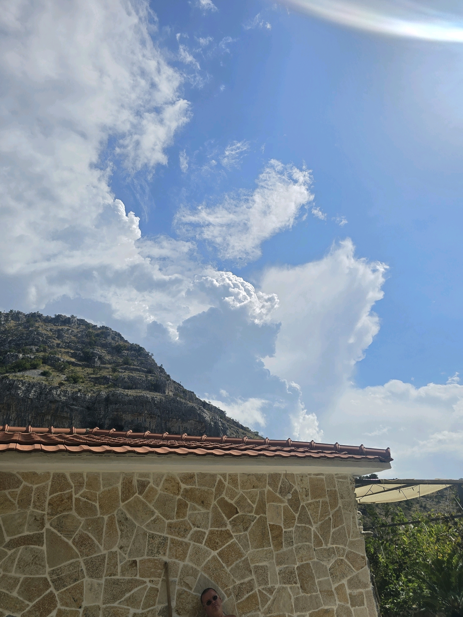 An outdoor shot captures a man in sunglasses leaning against a stone wall. He is wearing a light-colored t-shirt and his head is tilted back, looking towards the sky. Behind him, the stone wall is constructed with irregularly shaped, light-colored stones. Above the wall, a red tiled roof is visible, and beyond that, a steep, rocky mountain covered in sparse vegetation rises up to meet a dynamic sky filled with dramatic, white cumulus clouds against a bright blue background. The sun casts a bright glare in the upper right corner of the frame. The overall impression is a sunny, mountainous landscape with a stone building in the foreground. The scene is likely located in or around Podgorica, Montenegro, given the characteristic mountainous terrain and Mediterranean-style architecture.