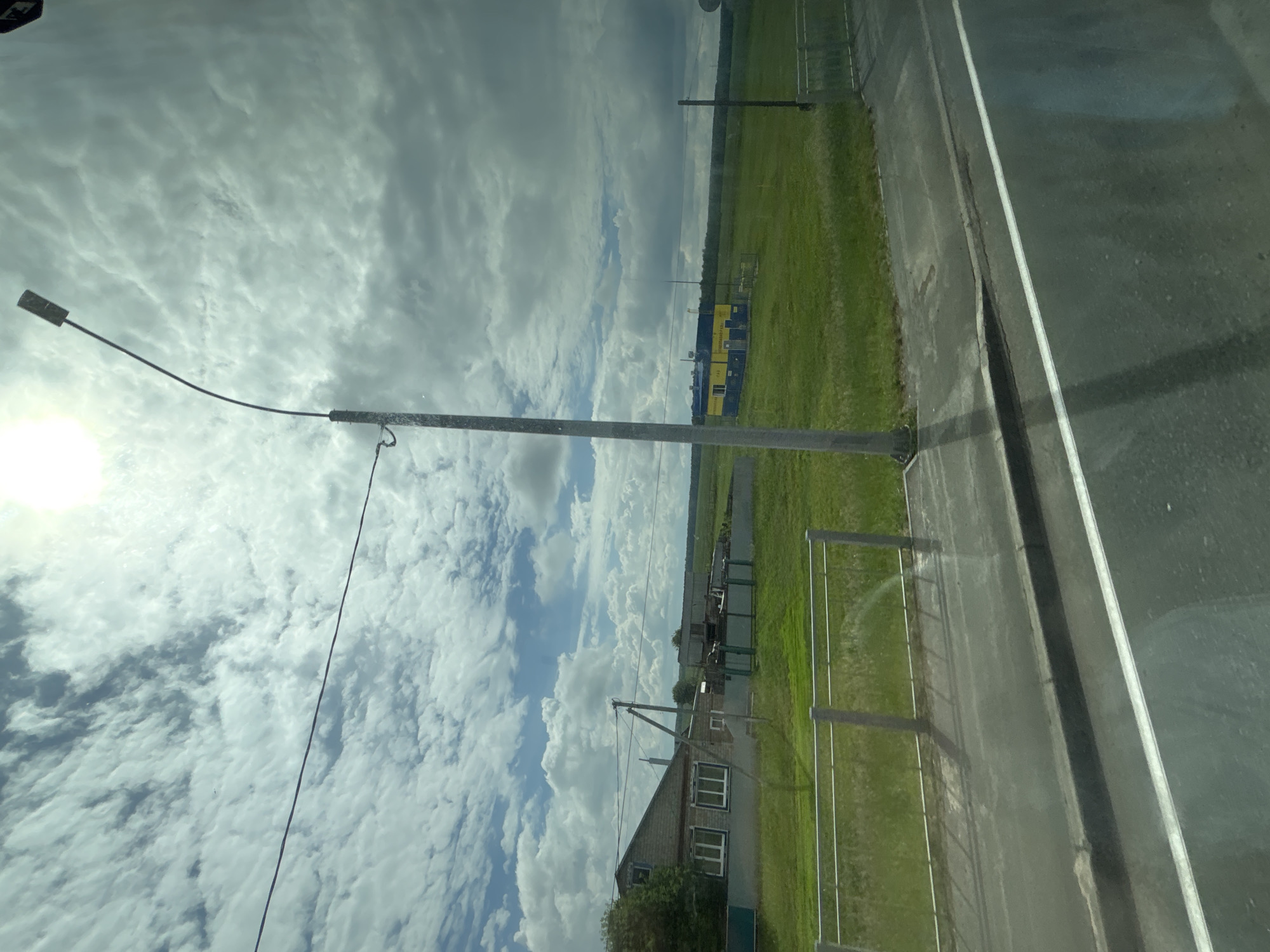 The image shows a view from a vehicle, looking out at a rural or semi-rural landscape under a cloudy sky with the sun shining through. In the foreground, there is a road with a white line, a curb, and a reflective surface. A silver metal fence runs along the edge of the road, separating it from a grassy field.
Further into the scene, there is a grey concrete lamppost with a curved arm extending upwards, topped by a dark lamp. A wire is attached to the lamppost and stretches horizontally across the sky. To the left of the lamppost, a house with a brick facade, light-colored window frames, and a grey roof is visible. Adjacent to the house is a dark green fence. In the middle ground, a long, low building with a yellow and blue facade stands in the grassy field. Beyond this building, a line of trees forms the horizon. The sky is filled with a mix of white and grey clouds, with bright sunlight filtering through from the upper left corner of the image. The scene appears to be in the vicinity of Лаяшты, Russia, given the architectural style and landscape. There are no people visible in the image.