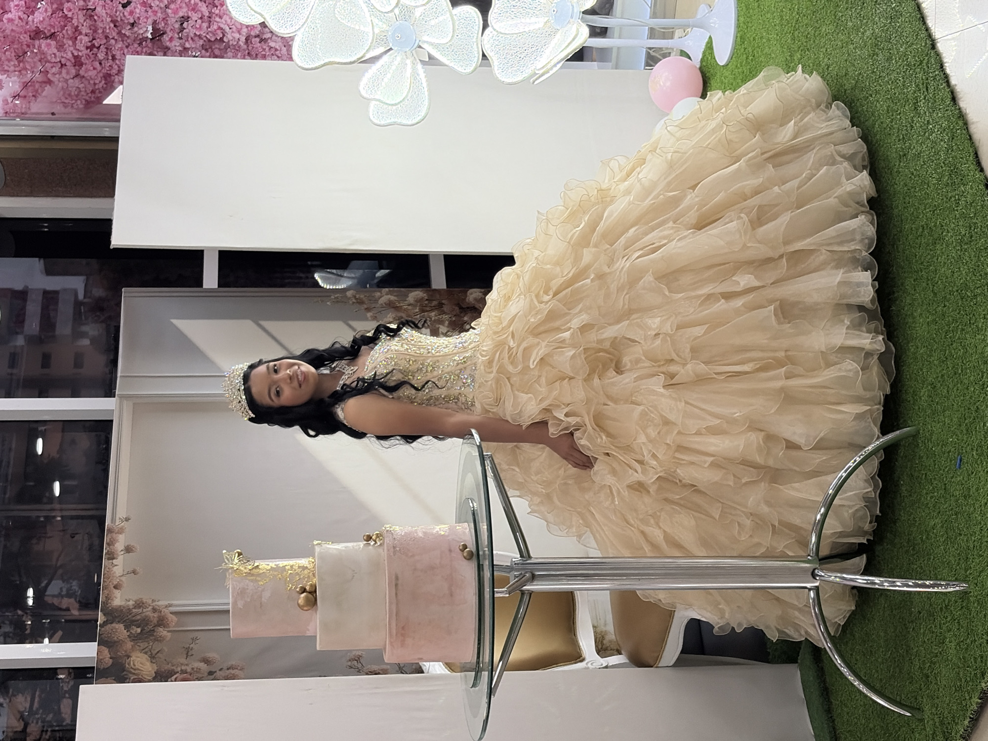 The image displays a young woman, likely a teenager celebrating a significant event such as a quinceañera, posing in an elaborately decorated indoor setting. She has long, dark, wavy hair and is wearing a sparkling tiara. Her dress is a voluminous, light champagne or beige-colored ball gown with a ruffled skirt and an embellished bodice. She is smiling broadly, conveying a sense of happiness and celebration.
To her left, on a modern glass-topped stand with a metallic base, sits a three-tiered cake. The cake features light pink and white tiers, adorned with gold leaf accents and small golden spherical decorations.
The background comprises several distinct elements. To the far left, there is a wall or backdrop covered in a dense pink floral or cherry blossom pattern. Behind the woman and the cake, a large, plain white panel or backdrop is visible. The floor beneath the woman and extending to the right is covered in artificial green turf. Overhead, delicate white, flower-shaped decorative lights are suspended, and a single pink balloon rests on the floor near a white platform. Reflections of what appear to be building windows can be seen in the background behind the cake. The overall scene suggests a festive and elegant event, consistent with a celebration in Ciudad Guayana, Venezuela. No text is visible in the image.