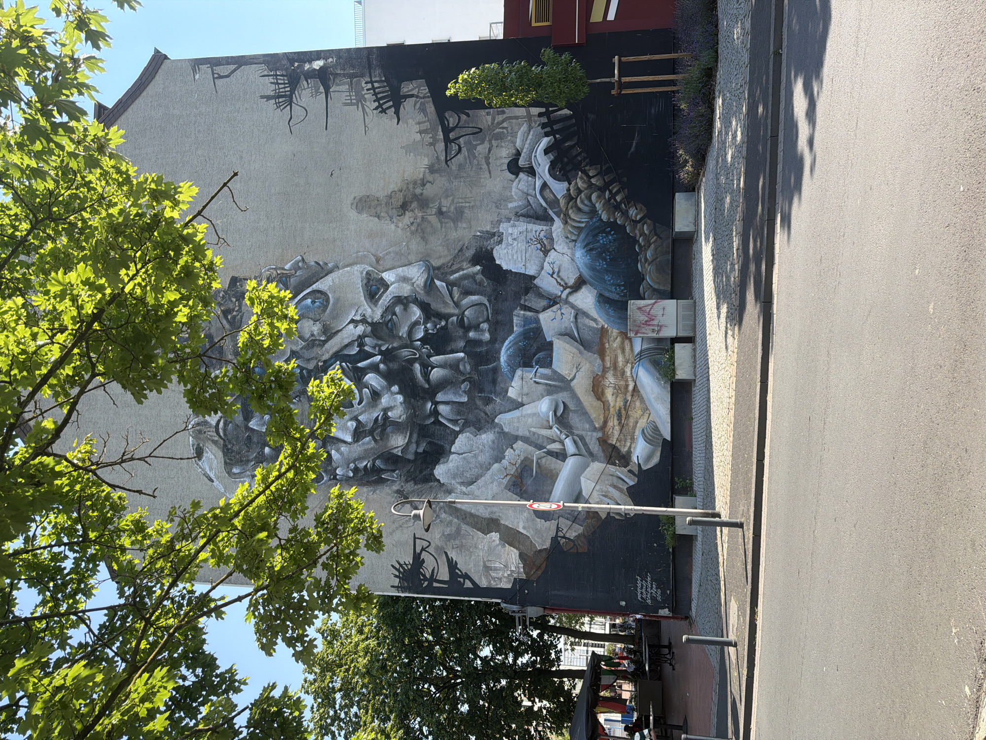 The image shows a large mural painted on the side of a building in an urban setting. The mural depicts a complex, surreal scene with abstract forms that appear to be faces or masks, rendered in shades of gray and white. There are also elements that suggest a landscape or industrial environment, with dark, jagged shapes and circular, bluish forms. The mural covers a significant portion of the building's facade, which is painted a light, textured color.
In the foreground, a street is visible with a gray asphalt surface and a concrete curb. Sidewalks are present on either side of the street, paved with cobblestones and concrete slabs. Several dark gray bollards are positioned along the sidewalk. A street lamp with a curved arm and a white lamp shade stands on the sidewalk in front of the mural. A red and white circular traffic sign is attached to the lamppost.
To the left of the mural, there are trees with lush green foliage, some of which cast shadows on the building and the sidewalk. Beyond the trees, a glimpse of a building with an awning and tables and chairs suggests an outdoor seating area for a cafe or restaurant.
To the right of the mural, a slender, young tree is planted in the ground, supported by wooden stakes. A patch of lavender flowers is visible behind the tree.
The sky is clear and blue, indicating a sunny day. The overall impression is of a vibrant urban environment with a striking piece of street art as the central focus.
