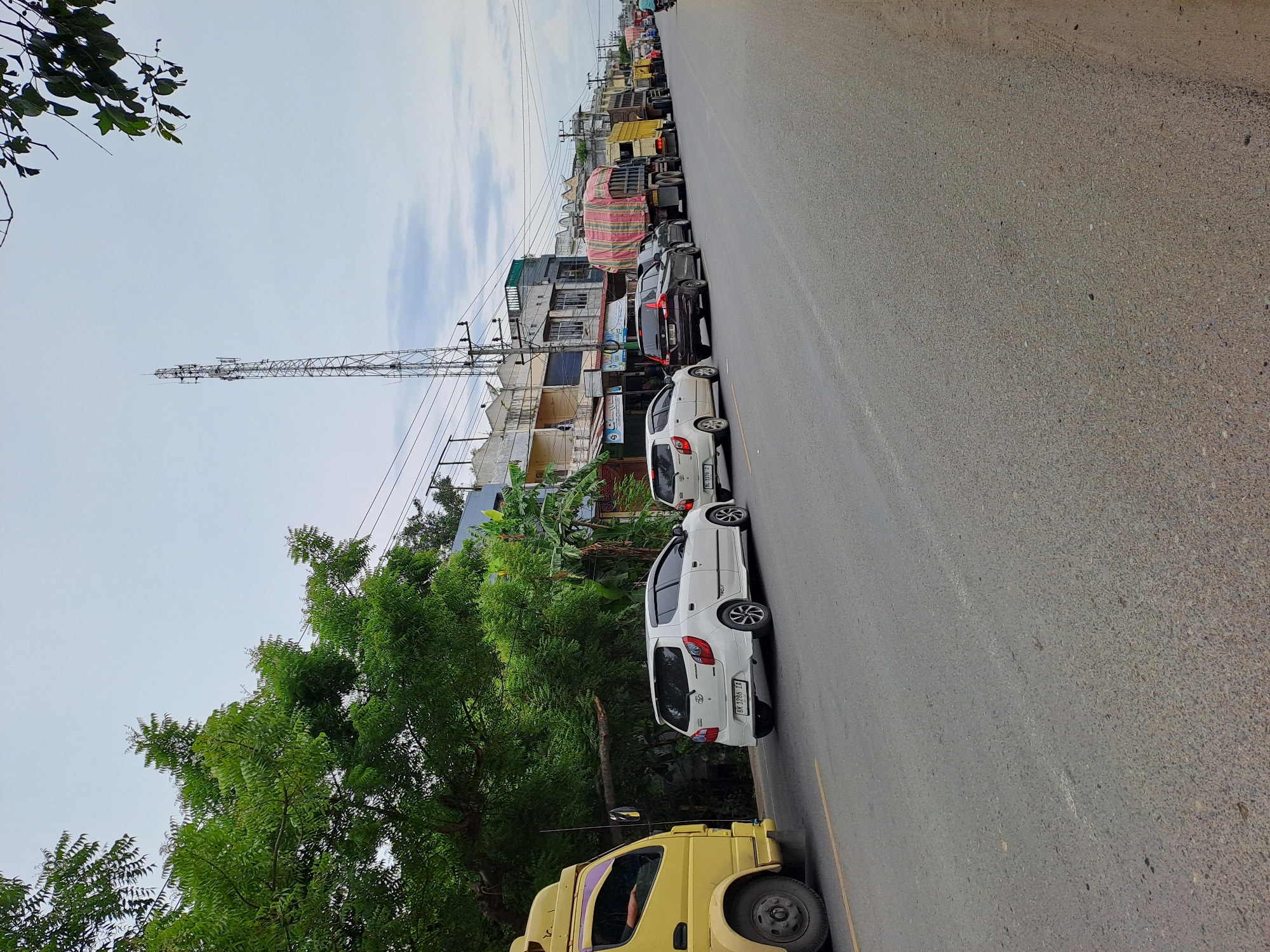 This is a street scene in Tingkeummanyang, Indonesia, with a long line of vehicles on a paved road under a cloudy sky. A yellow truck is partially visible on the left, facing away from the camera. Several cars, including a white hatchback and a black SUV, are stopped in traffic behind it. Further down the road, a series of large trucks, some covered with red and white tarpaulins, are also queued. Buildings with storefronts line the right side of the road, with a tall cellular tower visible in the distance. Lush green trees and foliage are present on the left side of the image, providing shade. The road appears to be a two-lane street with a yellow dividing line. The overall atmosphere suggests a traffic delay.