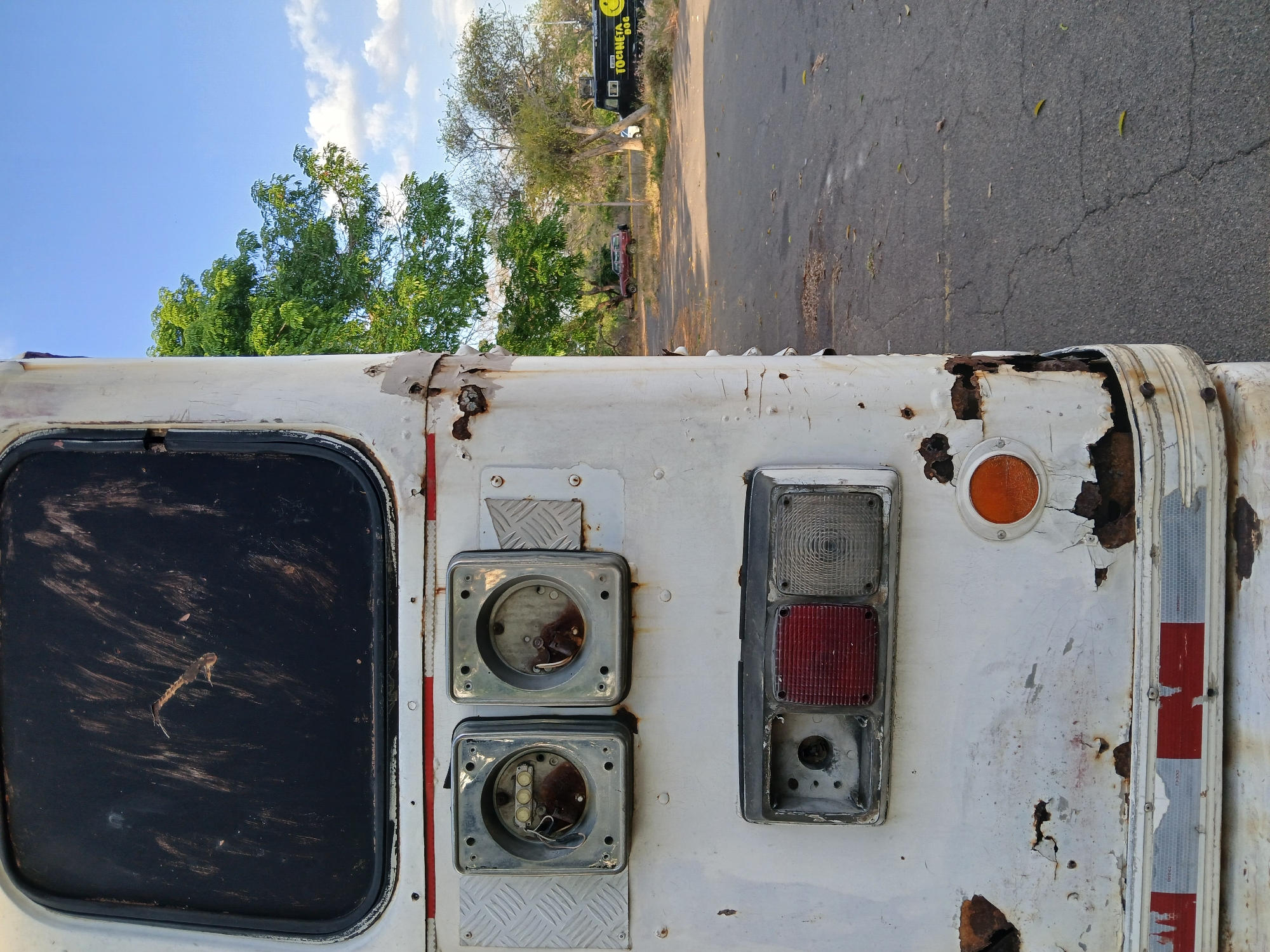 The image shows the rear of a rusted, white vehicle parked on a paved road in Ciudad Guayana, Venezuela. The vehicle appears to be abandoned or neglected, with significant rust and paint damage. The rear window is dirty. Several taillight assemblies are present, some missing lenses or bulbs. The surroundings include a street with trees on the side. A food truck is visible with a smiley face.
