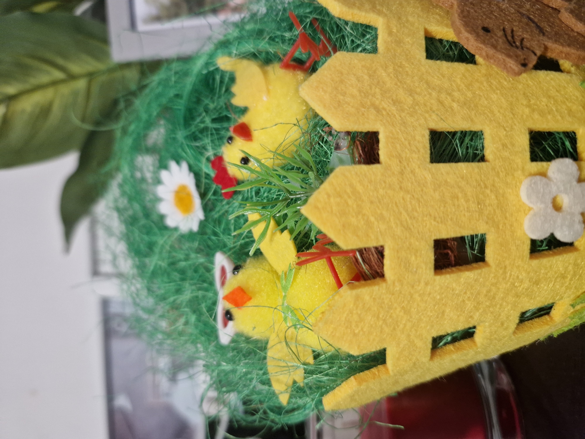 This image depicts a decorative Easter-themed arrangement located in the city of Unknown, Unknown. The primary subject is a yellow felt basket or fence-like structure with multiple square cutouts, occupying the lower and side portions of the frame. Inside this structure, vibrant green artificial grass is visible. Two small yellow chick figurines are positioned within the grass. The chick on the left faces slightly right, features an orange beak, black dot eyes, and wears white rectangular sunglasses. A small green bow is tied around its neck. The chick on the right faces left, has a red comb, a red beak, and black dot eyes. Both chicks have visible red plastic feet. A single white daisy with a yellow center is situated in the green grass towards the upper left of the chicks. Small sprigs of artificial green foliage are also dispersed within the grass. To the far left, a portion of a red candle in a clear glass container is visible. On the far right, a brown, textured figurine resembling a rabbit is partially visible behind the yellow felt. In the background, out of focus, a light-colored wall is present, with blurred hints of a large green plant on the right and dark rectangular objects, possibly frames or screens, on the left.