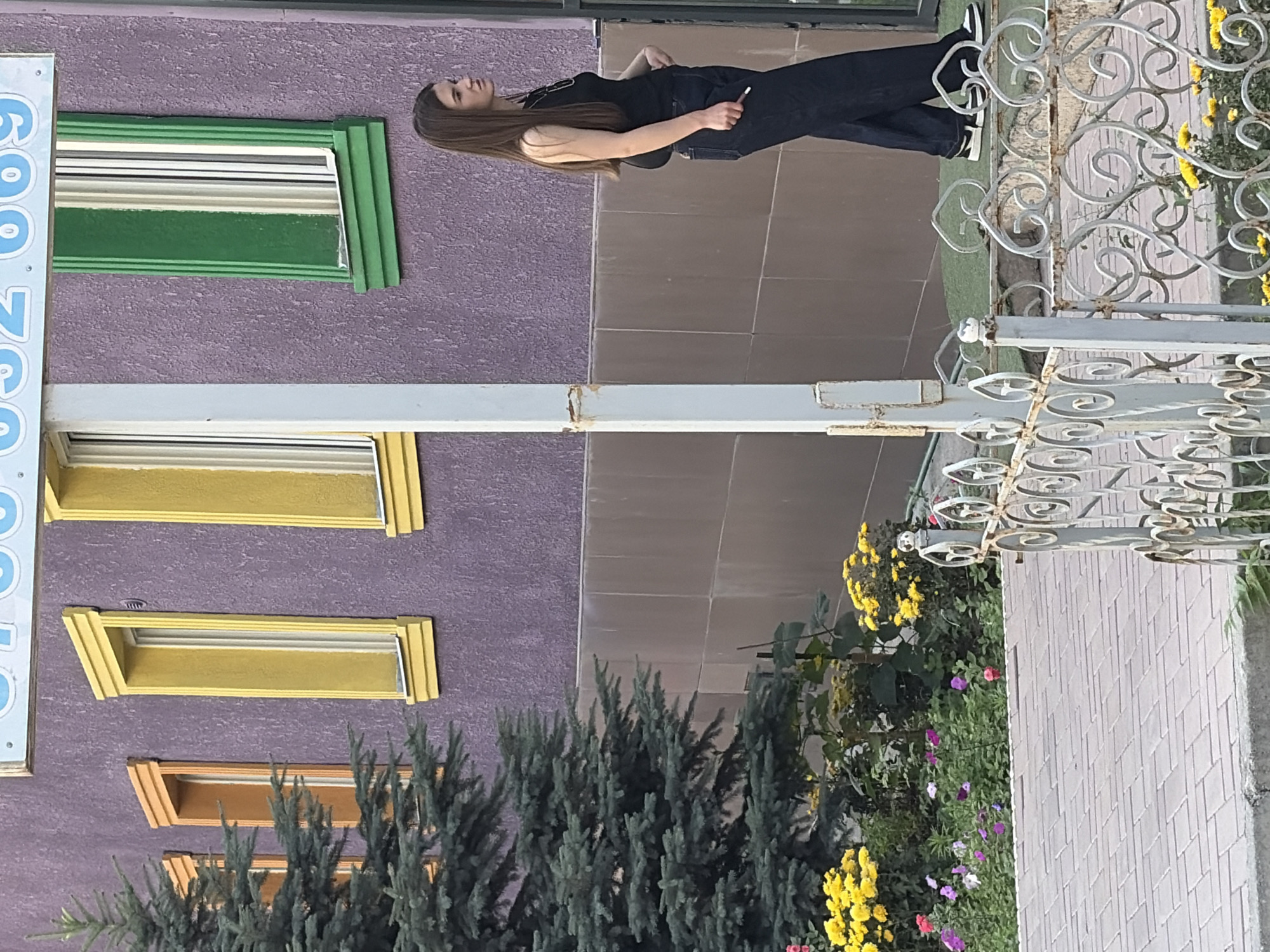 A young woman with long brown hair stands to the right of the frame, facing left and looking away. She is wearing a black tank top and dark, wide-leg pants. In her right hand, she holds a cigarette. She is positioned in front of a building with a textured purple facade, featuring brightly colored windows with orange, yellow, and green frames. To the left of the building, a large green evergreen tree stands tall. In the foreground, a flower bed with yellow and pink flowers is visible, separated from a paved walkway by a decorative white metal fence. The scene appears to be outdoors in an urban setting, possibly during the daytime.