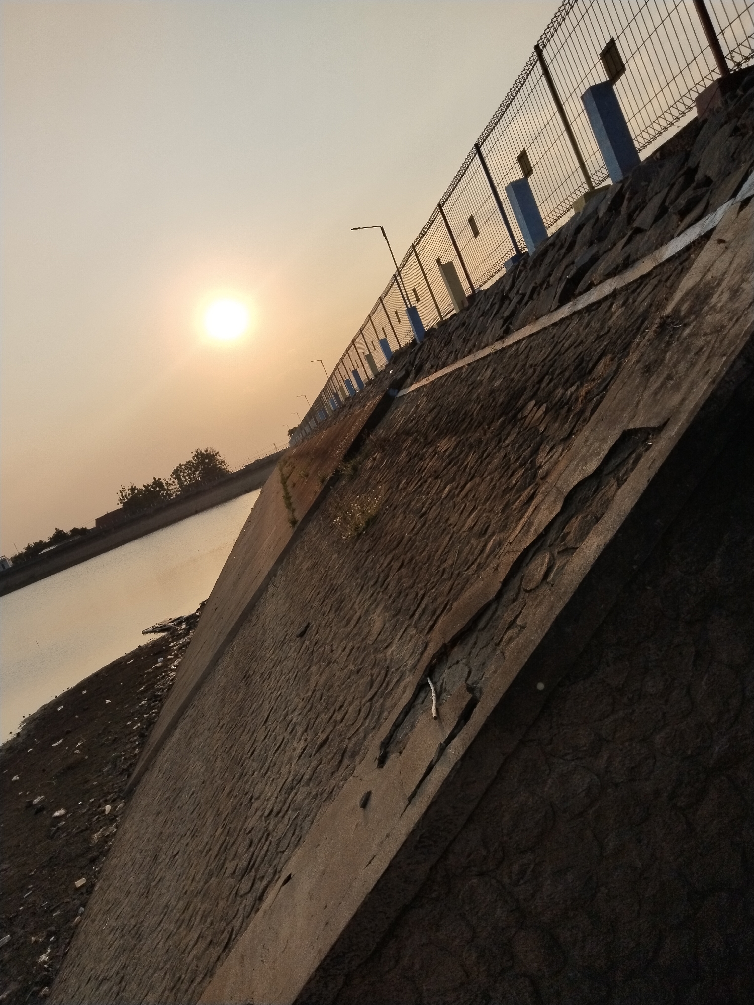 The image depicts a perspective view of a dam or embankment, angled upwards towards the sky. The sun is setting or rising, casting a warm, diffused light across the scene. The dam itself is constructed from dark, rough stones, with a concrete structure on its face. A metal fence runs along the top of the embankment, with blue and white posts. In the background, a body of water is visible, with trees and buildings on the opposite shore. The ground at the base of the dam appears to be composed of dark soil and debris. The image appears to be taken in Blimbing, Indonesia.