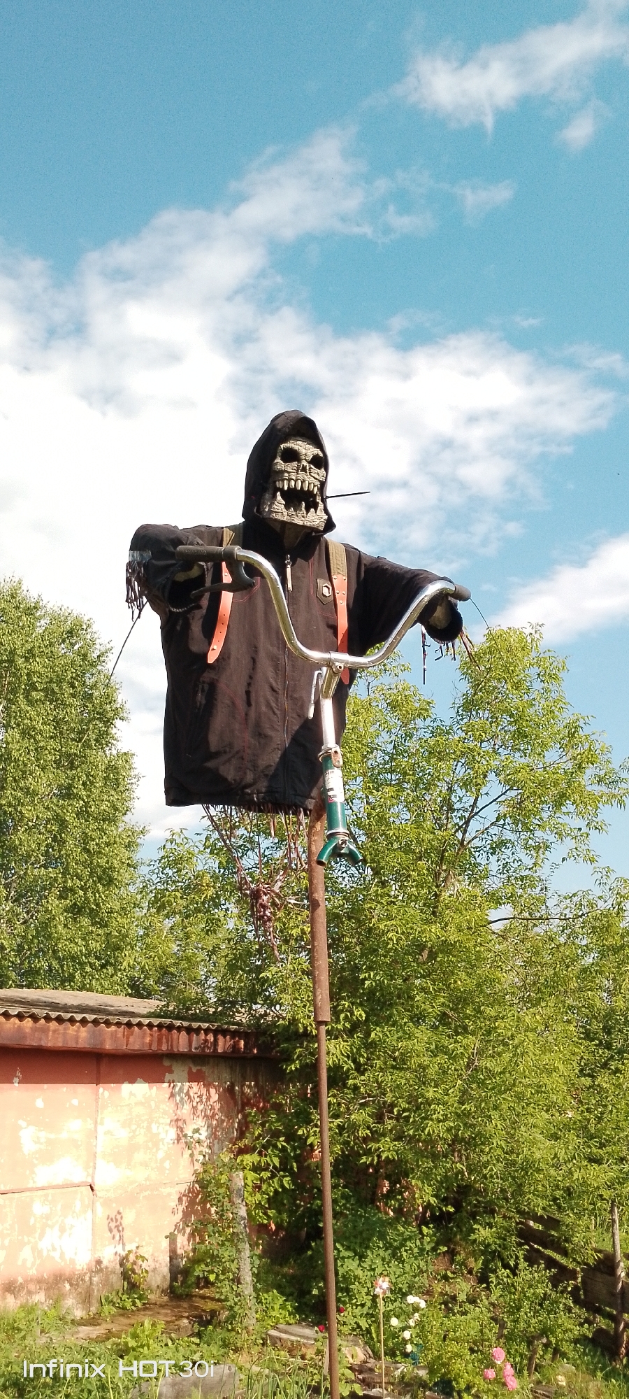A grim reaper figure, possibly a scarecrow, is mounted on a metal pole in an outdoor setting. The figure wears a black hooded robe and a skull mask. It is posed as if riding a bicycle, with its arms on a chrome handlebar. The background consists of lush green trees and a blue sky with white clouds. A dilapidated orange-and-white building is visible to the left, partially obscured by greenery. The scene appears to be in a rural or semi-rural area.