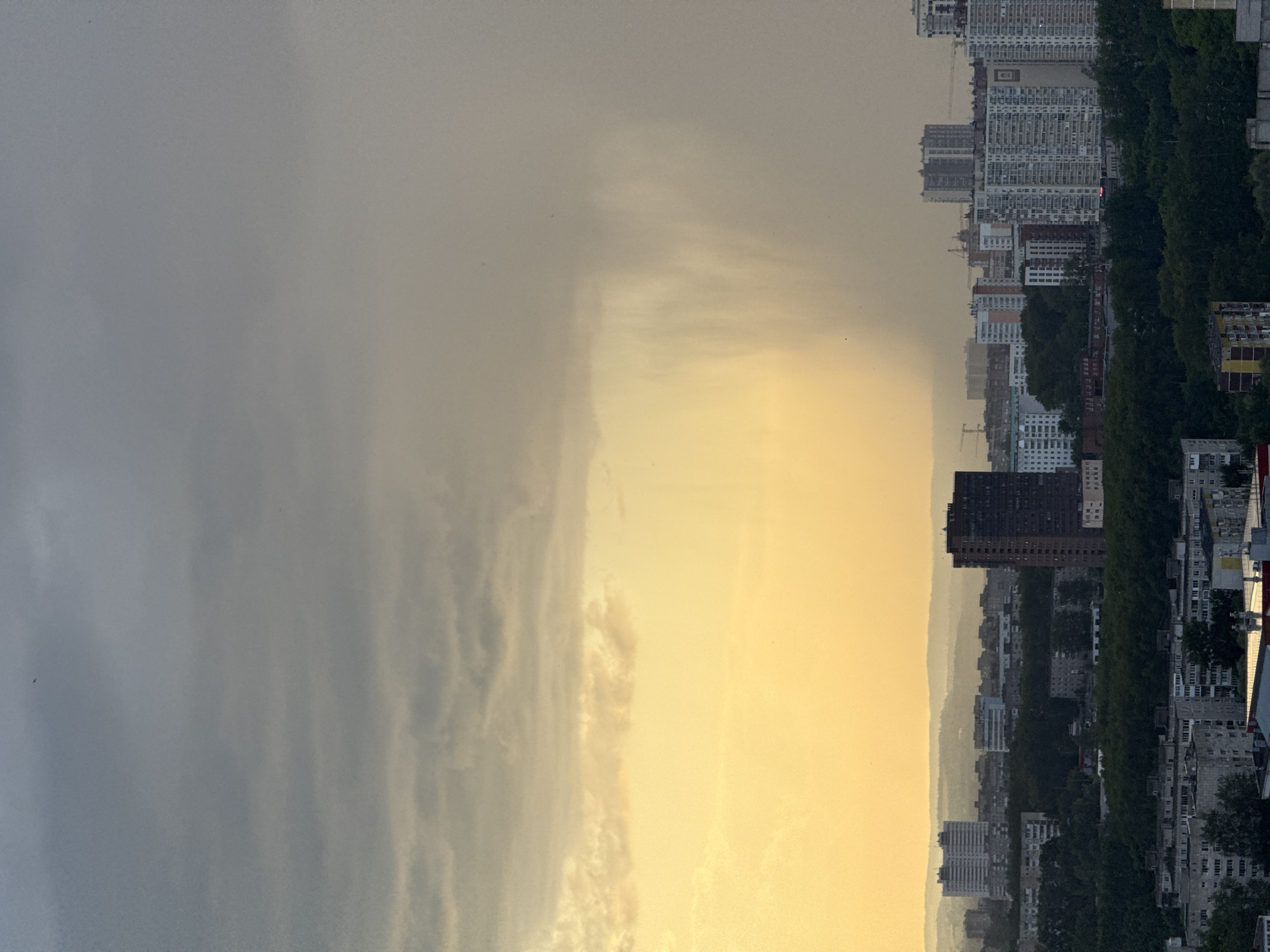 The image depicts an urban landscape in Shirkovo, Russia, under a dramatic sky. The foreground is dominated by residential buildings and dense green trees. In the mid-ground, more buildings of varying heights are visible, including a prominent tall, dark-colored apartment building. The background shows a hazy cityscape with more distant buildings and rolling hills. A striking feature is a heavy rain shower occurring in the distance, illuminated by the warm, golden light of the setting sun on the left side of the frame. The sky above is a gradient of gray and yellow, with dark clouds on the left transitioning to a lighter, overcast sky on the right. No people are clearly visible in the image. The overall atmosphere is one of an impending or ongoing storm over a city.