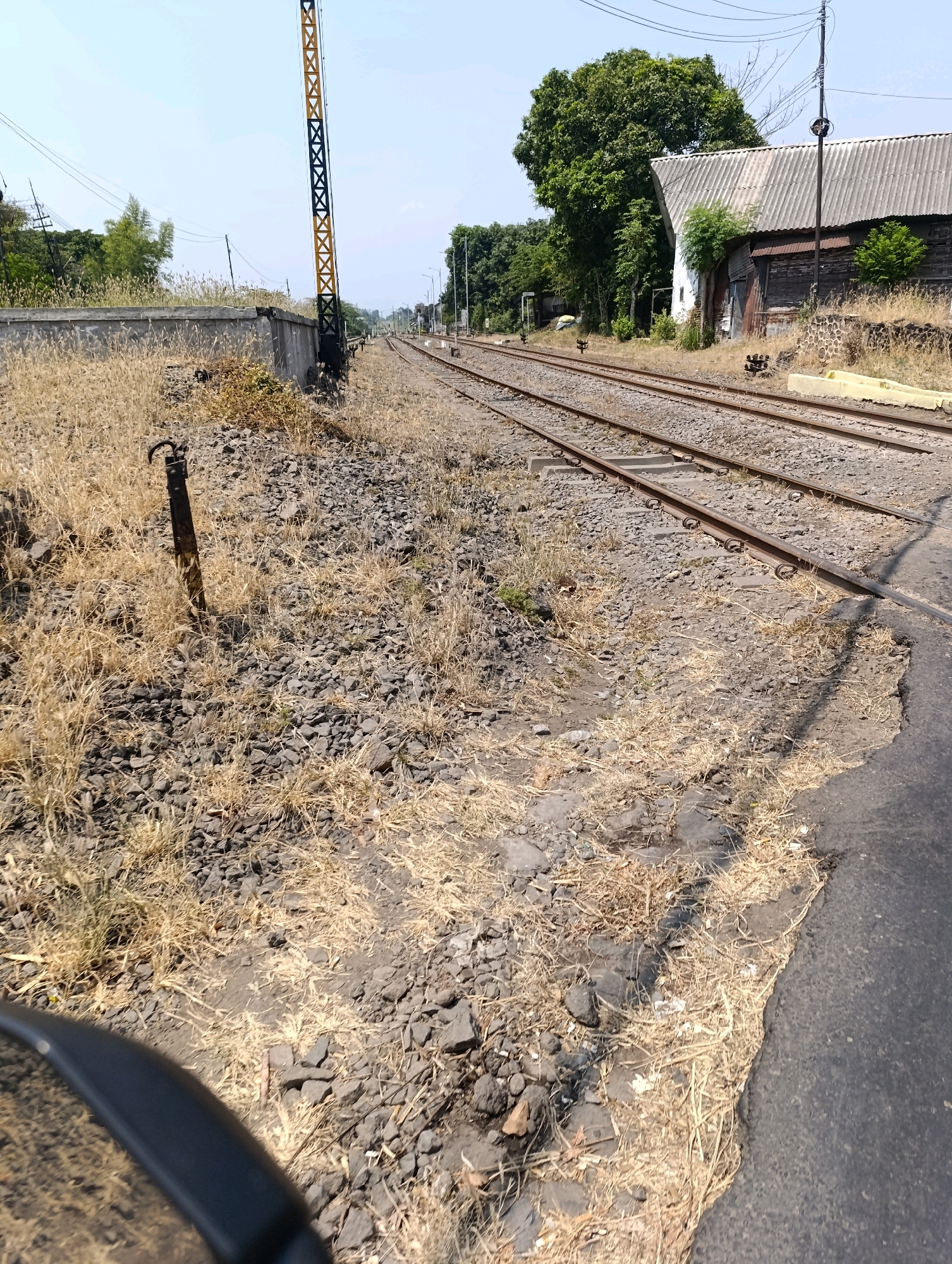 The image shows a railroad track in Glagah Sari, Indonesia, on a sunny day. The tracks are surrounded by dry grass and gravel. A large tree is visible in the background, along with a building that appears to be a warehouse or storage facility. There is also a signal pole with yellow and black markings. The foreground of the image shows a portion of what appears to be a black vehicle, possibly a motorcycle, based on the curved edge visible. No people are present in the image.