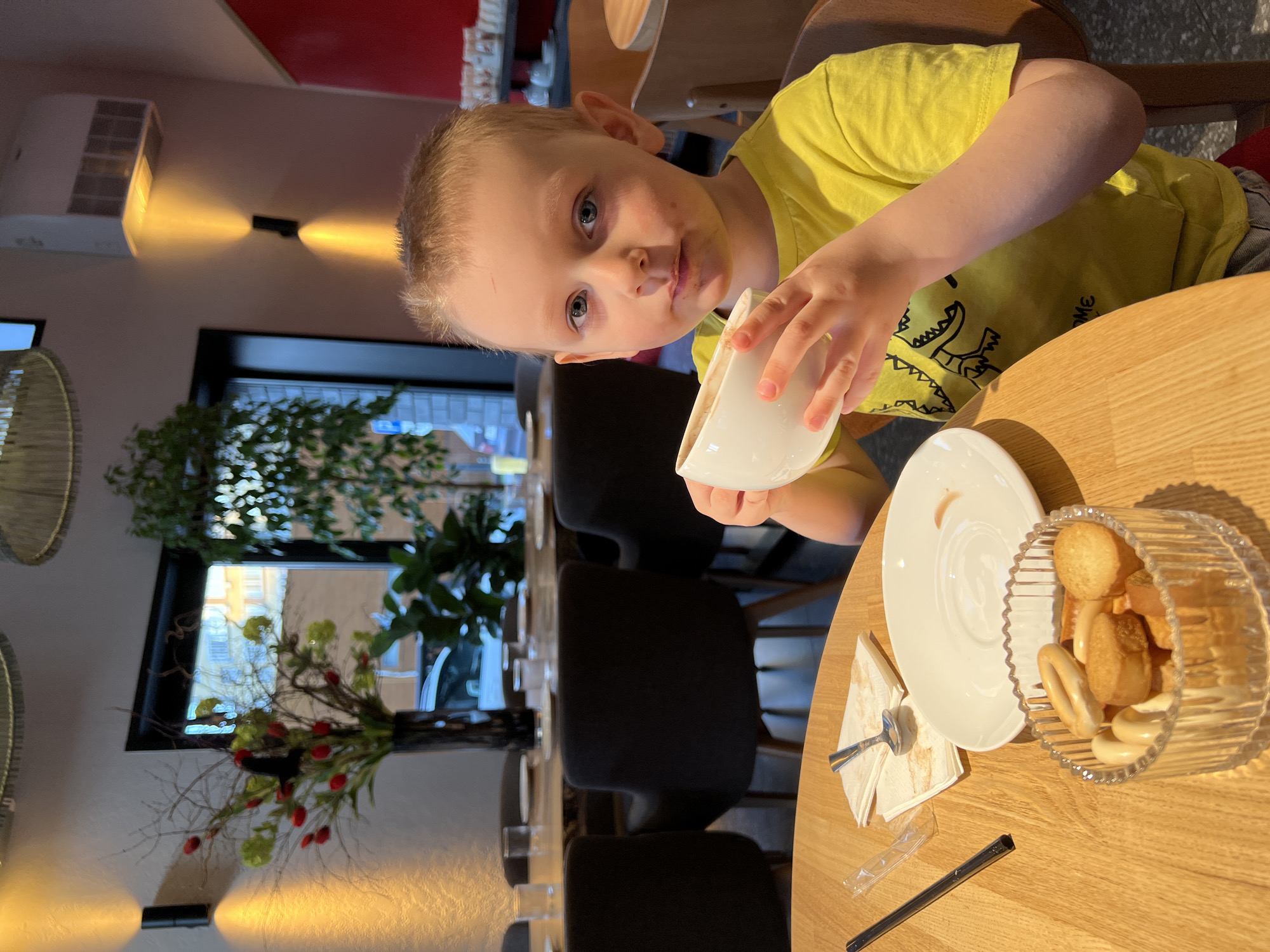 A young boy in a yellow T-shirt is drinking from a white cup at a table in an indoor setting, likely a cafe or restaurant. The table is made of light-colored wood and has a white saucer, a glass bowl of small baked goods (possibly cookies or crackers), a napkin, a spoon, and a straw. In the background, there are black chairs, a window with greenery visible outside, and interior lighting fixtures. The location is Mytishchi, Russia.