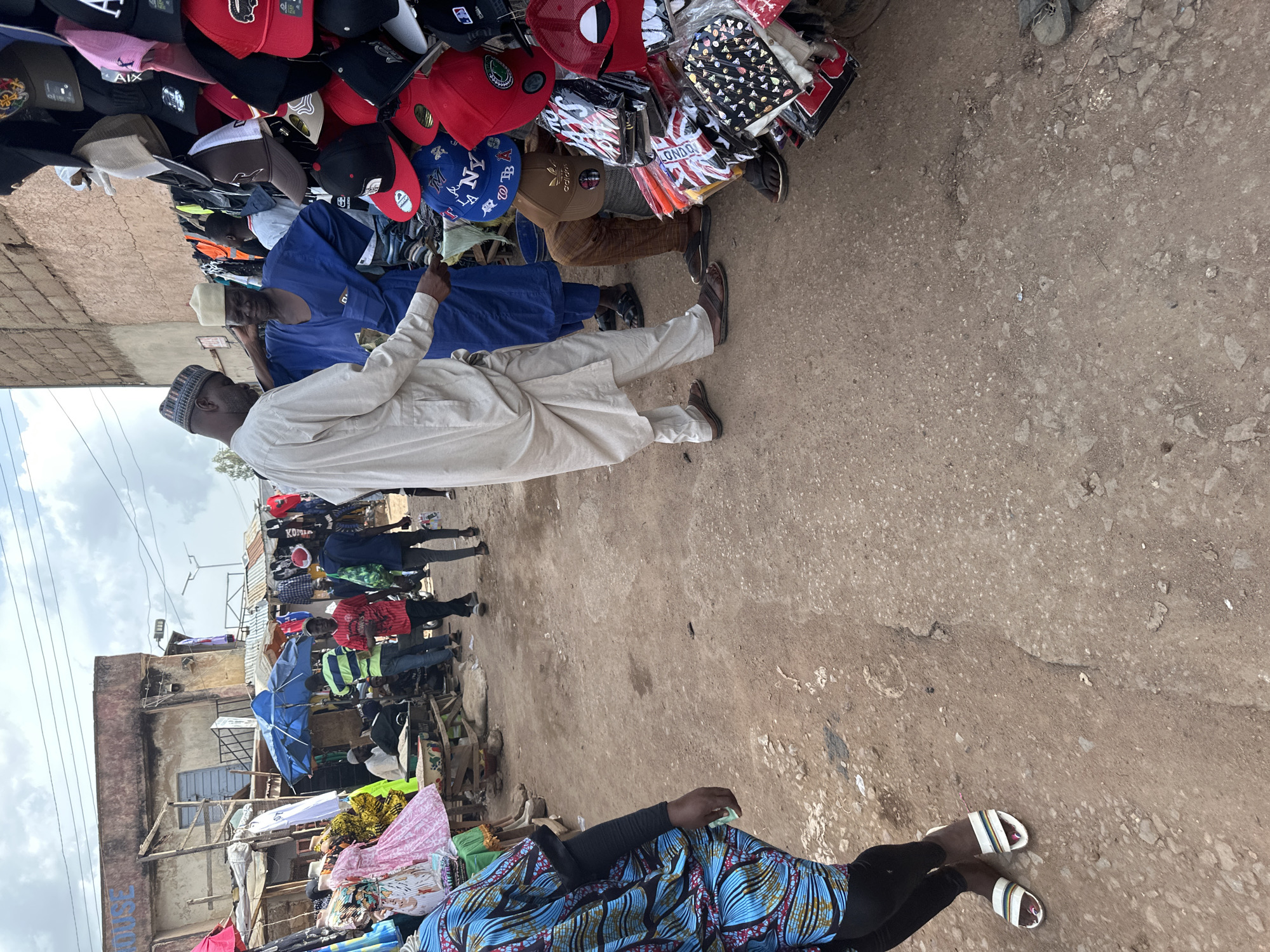 In Bukuru, Nigeria, a bustling street market is captured. The scene depicts a variety of people engaged in commerce. In the foreground, a person is seen from the waist down, wearing a colorful patterned outfit and striped sandals. To their left, a man in a light-colored kaftan and cap converses with another man in a blue traditional outfit, who is seated. Behind them, a stall displays a wide array of baseball caps and other headwear. Further in the background, other market-goers are visible, some browsing through clothing and accessories. The market appears to be set up along a dusty, unpaved street, with various structures and stalls lining the sides. The overall atmosphere suggests a lively commercial environment.