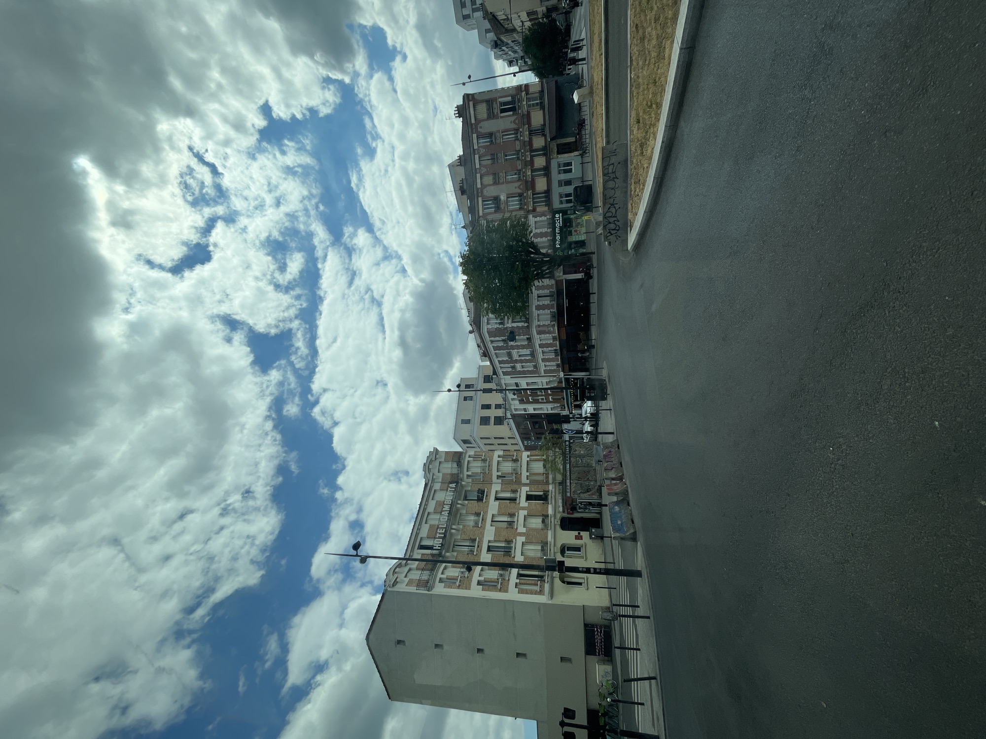 This is a wide-angle, exterior shot of a street scene in Ivry-sur-Seine, France. The perspective is from a low vantage point, looking upwards at a diagonal. The sky dominates the left half of the frame, filled with dramatic, fluffy white clouds interspersed with patches of bright blue. The right half of the image shows a paved street, a sidewalk, and several multi-story buildings lining the street.
The buildings are of various architectural styles, appearing to be residential or mixed-use. One building in the mid-ground has visible text that reads "HOTEL BENVENIDA," suggesting it is a hotel. Another building to its right has a sign that says "Pharmacie." The buildings have numerous windows, some with balconies. Trees are visible growing between the buildings.
The street is empty of people and cars, giving the scene a quiet, perhaps early morning or late evening feel. The pavement appears well-maintained, and there is a grassy area with a concrete curb alongside the street. Some graffiti is visible on the lower parts of the buildings and on some street furniture.
The overall impression is of an urban environment on a cloudy day, possibly mid-day given the brightness of the sky in some areas. The lighting suggests natural daylight. There is no indication of any specific event taking place; it appears to be a standard street view. The weather seems to be partly cloudy.