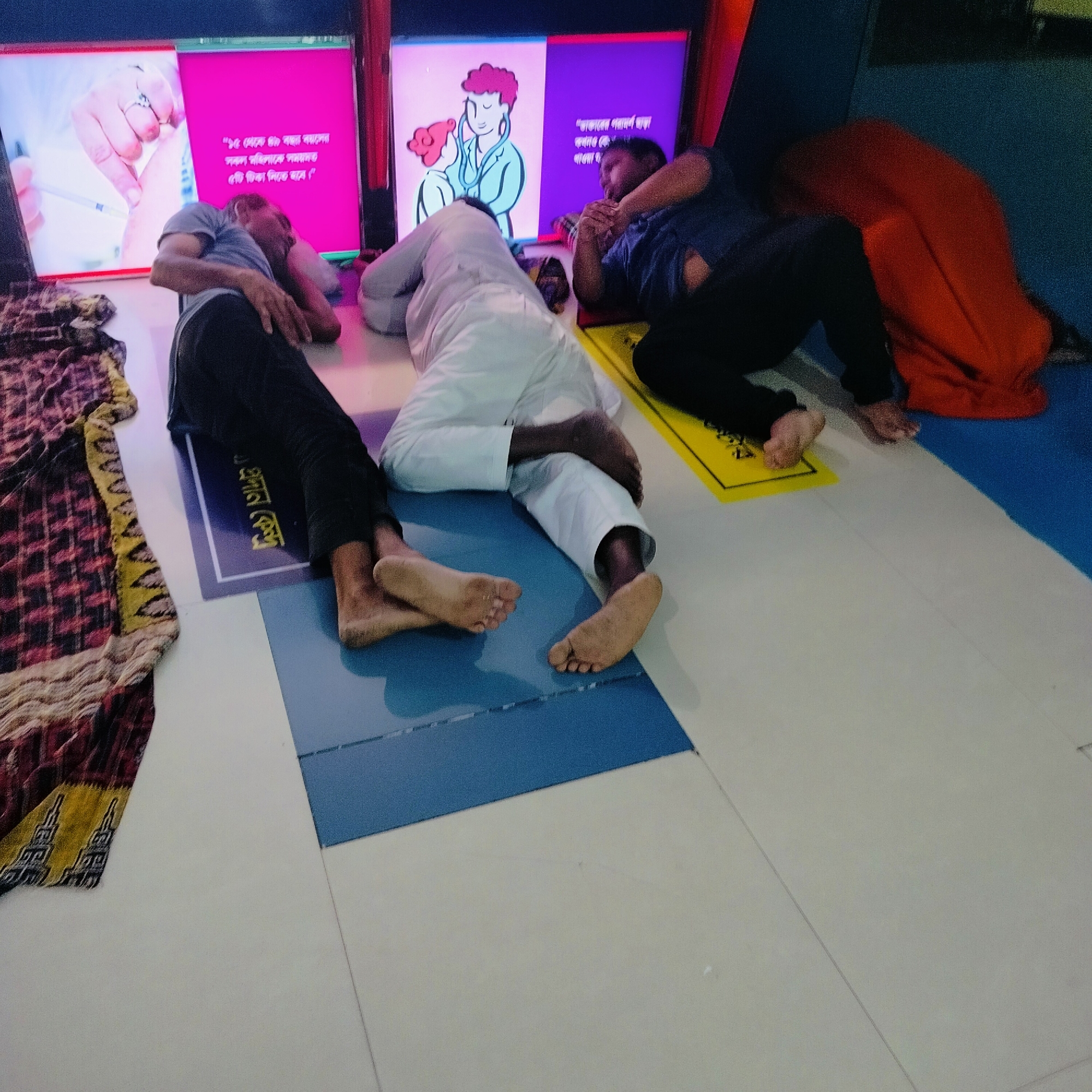 Three men are reclining on yoga mats in front of illuminated signs, presumably sleeping or resting, in what appears to be a public space. The men are of Asian descent. The first man is wearing a gray shirt and black pants, the second is wearing all-white clothing, and the third is wearing a dark blue shirt and black pants. The men are lying on individual yoga mats of different colors: purple, blue, and yellow. The mats are placed on a tiled floor. Beside the first man's mat, there is a folded, patterned fabric. Behind the men, there are three illuminated signs with text and images, suggesting the location may be a health center or public health campaign in Dhaka, Bangladesh, given the Bangla script present on the signs. One sign has an image of a hand with a syringe, another has a drawing of a doctor with a stethoscope, and the third is partially obscured but shows a person. The overall impression is of a waiting area or a temporary resting place.