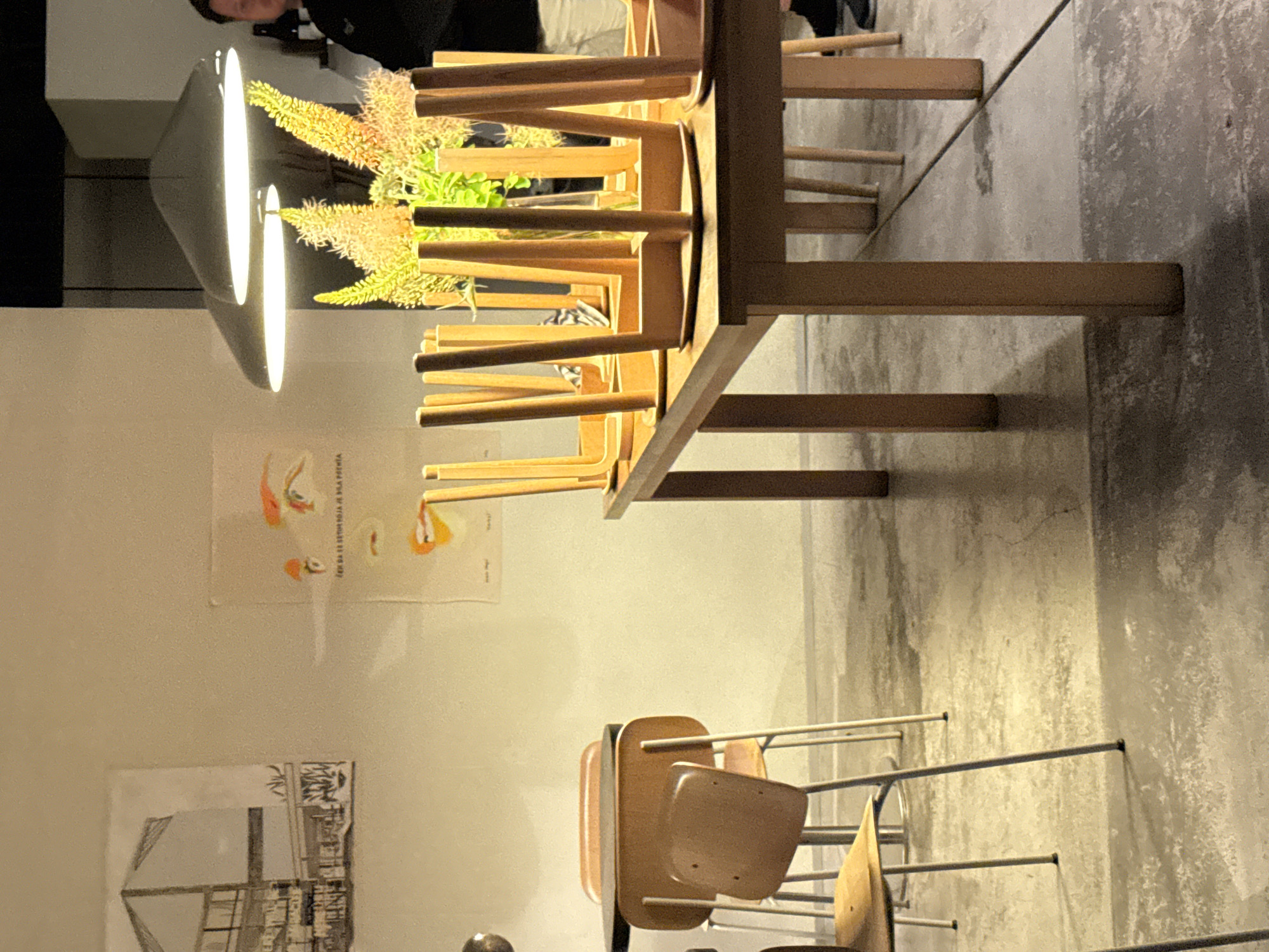 The image depicts the interior of what appears to be a restaurant or cafe, possibly in Belgrade, Serbia, with a minimalist and modern aesthetic. The scene is lit by ambient lighting from overhead pendant lamps, casting a warm glow. In the foreground, a wooden table is laden with numerous inverted wooden chairs, their legs pointing upwards, creating a geometric pattern. The table itself is simple and rectangular.
To the left, another table with a dark top is visible, accompanied by chairs. One chair, with a light brown wooden seat and back, is prominently featured in the mid-ground. Further to the left, a framed architectural drawing or poster hangs on the wall. A second poster, featuring a stylized illustration of eyes and lips, is also on the wall.
In the background, a person wearing a black top is partially visible, standing behind a table. A vase containing yellow and green floral arrangements is placed on the table in the mid-ground, adding a touch of color and organic form to the composition. The floor is made of concrete, showing some signs of wear and tear, contributing to the industrial-chic ambiance. The overall impression is one of a contemporary dining or gathering space, possibly closed for the evening given the stacked chairs.