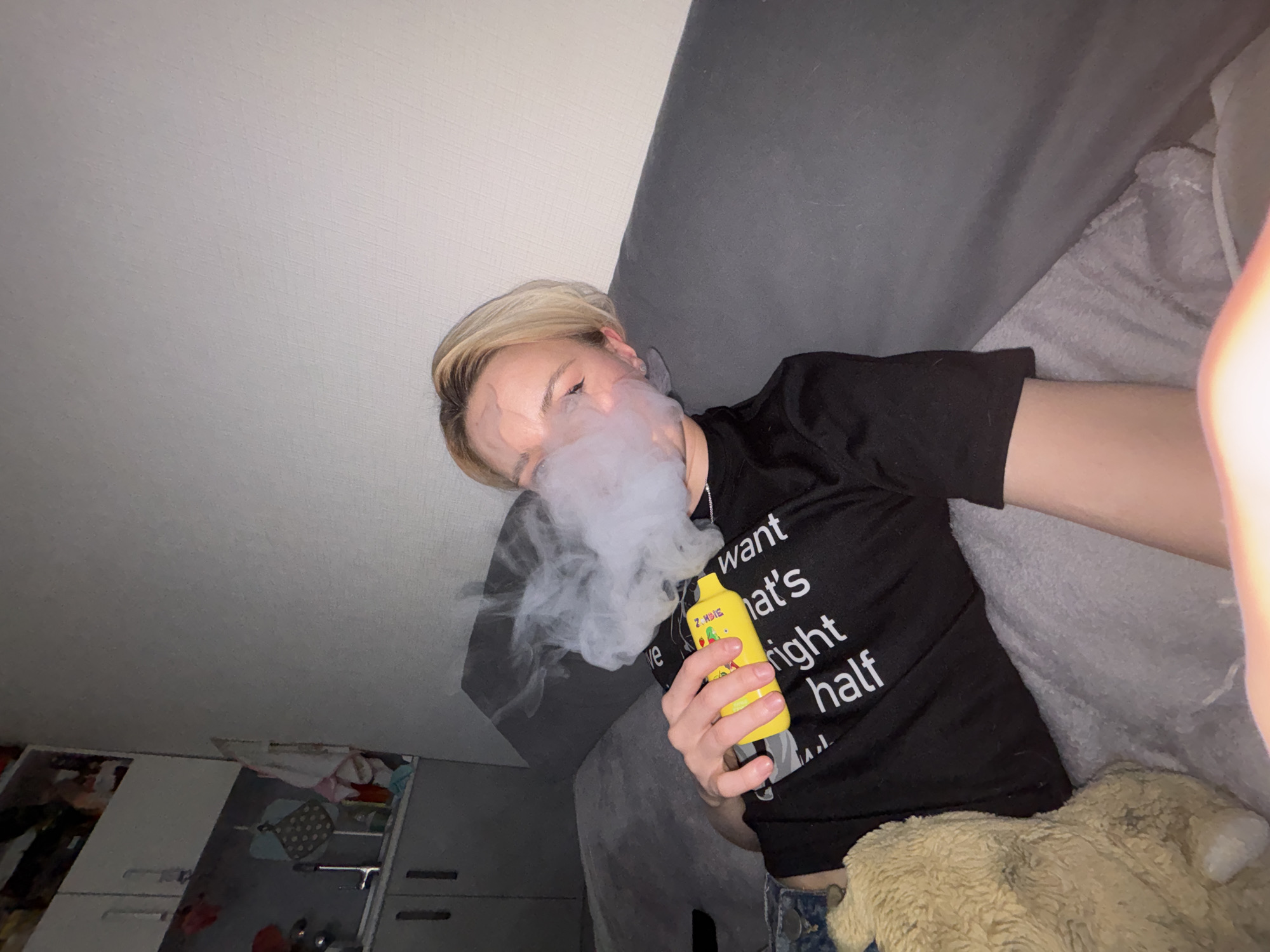 An individual with short, light-colored hair is depicted in a slightly angled, indoor shot, exhaling a large cloud of white vapor from a yellow vaping device. The person is wearing a black short-sleeved t-shirt with partially visible white English text on the front, which includes the words "want", "what's", "right", and "half". The yellow vaping device, held in their right hand, clearly displays the brand name "ZOMBIE" along with small, colorful fruit illustrations.
The person is reclining on what appears to be a grey couch or bed, with a fluffy brown blanket or plush item partially visible to their right. Their face is partially obscured by the exhaled vapor, and their eyes are either closed or cast downwards. The background reveals a simple room setting, featuring light-colored walls and ceiling, and parts of white or light-grey cabinets or furniture with drawers, suggesting a bedroom or living area. The lighting appears to be artificial, casting some shadows and creating a bright flare on the right side of the image. The overall scene suggests a casual and relaxed activity.
Location: Unknown, Russia.