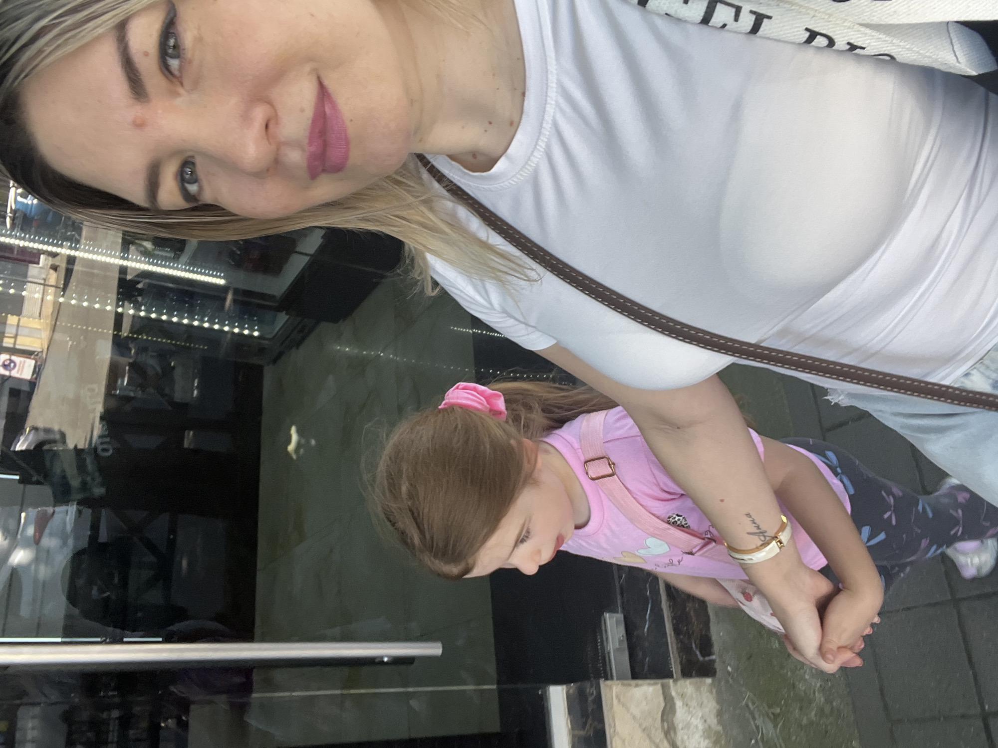 An image, rotated 90 degrees counter-clockwise, depicts a woman and a young girl. The woman, positioned in the upper portion of the frame, has blonde hair, light eyes, and pinkish-red lipstick. A small blemish is visible on her forehead. She wears a white t-shirt and a dark brown strap of a bag across her shoulder. Her right forearm bears a script tattoo, which appears to read "Shuna."
Below her, a young girl with light brown hair, tied in a high ponytail with a pink scrunchie, is visible. She wears a pink t-shirt with heart and animal print designs, dark patterned leggings (possibly with dragonflies), and a pink shoulder bag. The woman is holding the girl's hand, and the girl is looking downwards.
The setting appears to be an indoor or covered outdoor area, possibly a storefront, indicated by reflections in a large glass surface behind them. These reflections show strip lighting and indistinct shapes. The ground is a mix of light and dark surfaces. The woman has a soft smile, while the girl's expression is not clear as her face is mostly turned away. The partially visible text on the woman's shoulder bag strap shows "...T D...". The time of day appears to be daylight or well-lit indoors. The location is specified as Sochi, Russia.