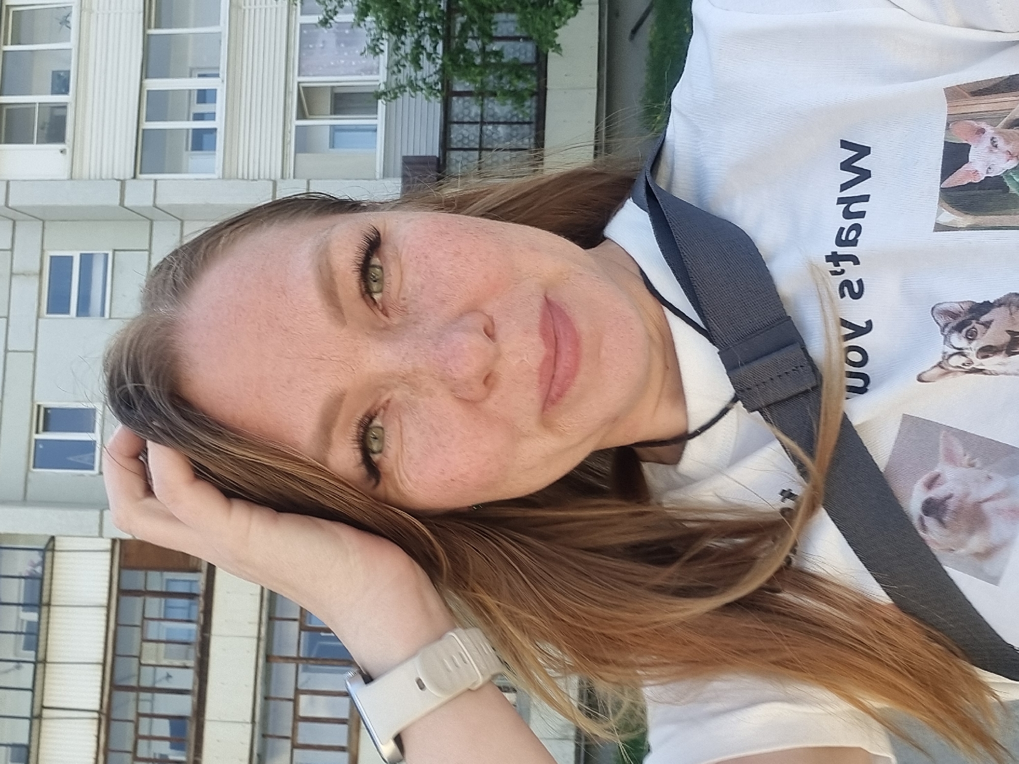 A woman is posing for a photograph. She is wearing a white T-shirt with cat images. Her hair is reddish-blonde and she has freckles. She is wearing a white smartwatch on her left wrist. The background features a building with multiple windows, suggesting an urban environment. Based on the prompt, the location is Chaikovsky, Russia.