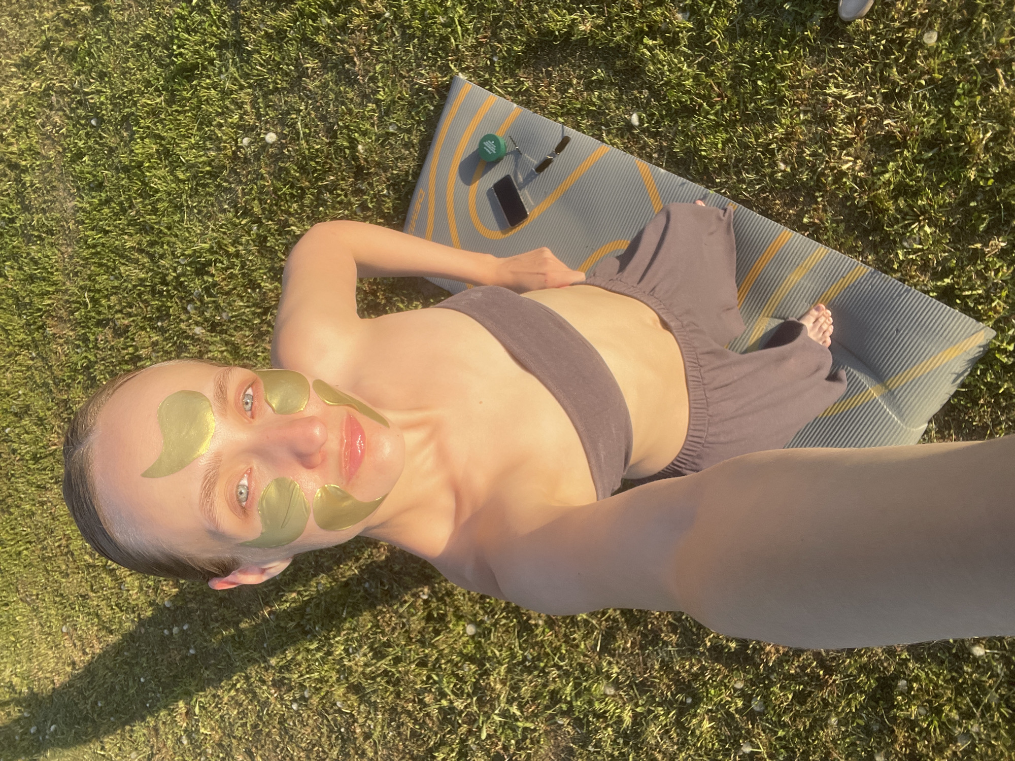 A fair-skinned woman with blonde hair pulled back lies on a yoga mat on a grassy lawn. She wears a brown sports bra and loose-fitting brown pants. Gold eye patches are applied to her face. Next to her on the mat are a green jar, sunglasses, and a smartphone. The image appears to be taken from a selfie angle, with the woman holding the camera. The location is possibly Подшивалово, Russia, though this cannot be forensically determined from the image alone.