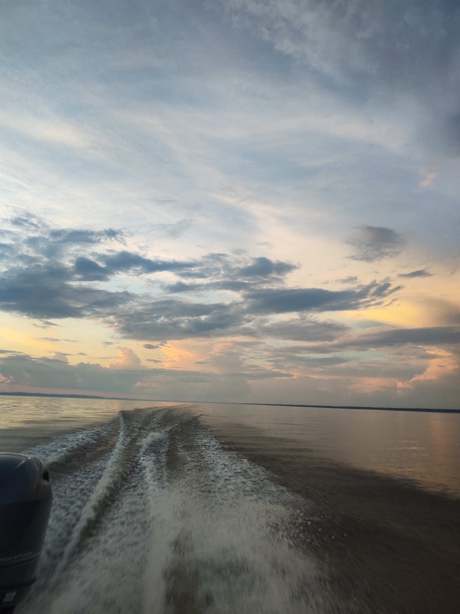 A wide shot captures the wake of a boat moving through calm water under a twilight sky. The sky is filled with wispy clouds in shades of blue, gray, and pink, with a warm glow of yellow and orange on the horizon. The water is dark and reflects the muted colors of the sky. In the foreground, the outboard motor of the boat is partially visible on the left, and the white foamy wake spreads out behind the vessel, creating a dynamic texture on the water's surface. The horizon line is visible in the distance, separating the water from the sky. The scene suggests a peaceful journey on a body of water at dusk. There are no people visible in the image. The location is not specified as being in the city of Галево, Russia.