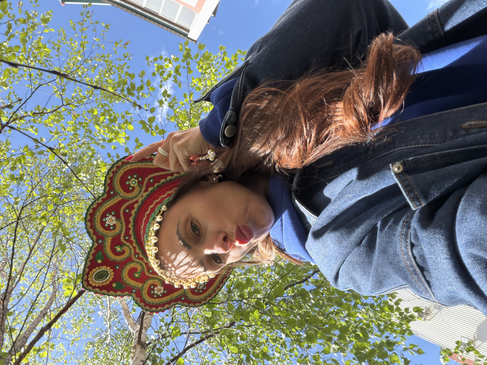 A low-angle shot shows a woman wearing a traditional Russian kokoshnik headdress. The headdress is red with gold embroidery and pearl embellishments. The woman is also wearing a blue sweatshirt and a denim jacket. Her hair is reddish-brown. She is looking down at the camera with a slightly puckered expression. Above her, green leaves of trees fill much of the frame, against a blue sky with some white clouds. Part of a building with white siding is visible in the upper right corner. The photograph may have been taken outdoors. Since there are no indications of where this photo was taken, I am unable to verify the location in the city of Birobidzhan, Russia.